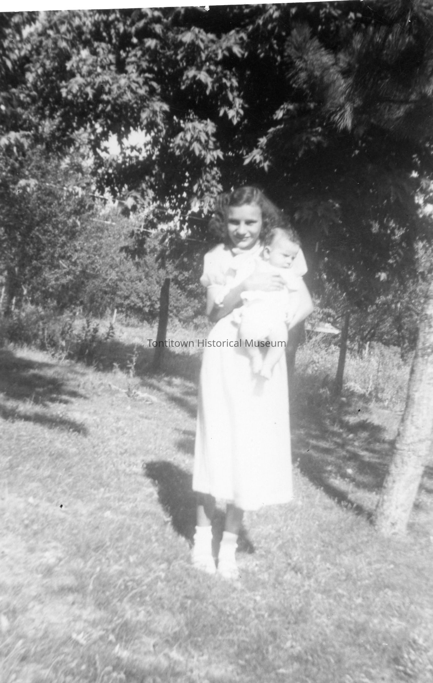 In this black-and-white photograph, a woman is lovingly holding a baby in her arms, standing outdoors amidst a serene natural setting with lush trees casting dappled shadows on the ground. The woman is dressed in a light-colored, ankle-length dress, complementing the bright and sunny ambiance of the garden.