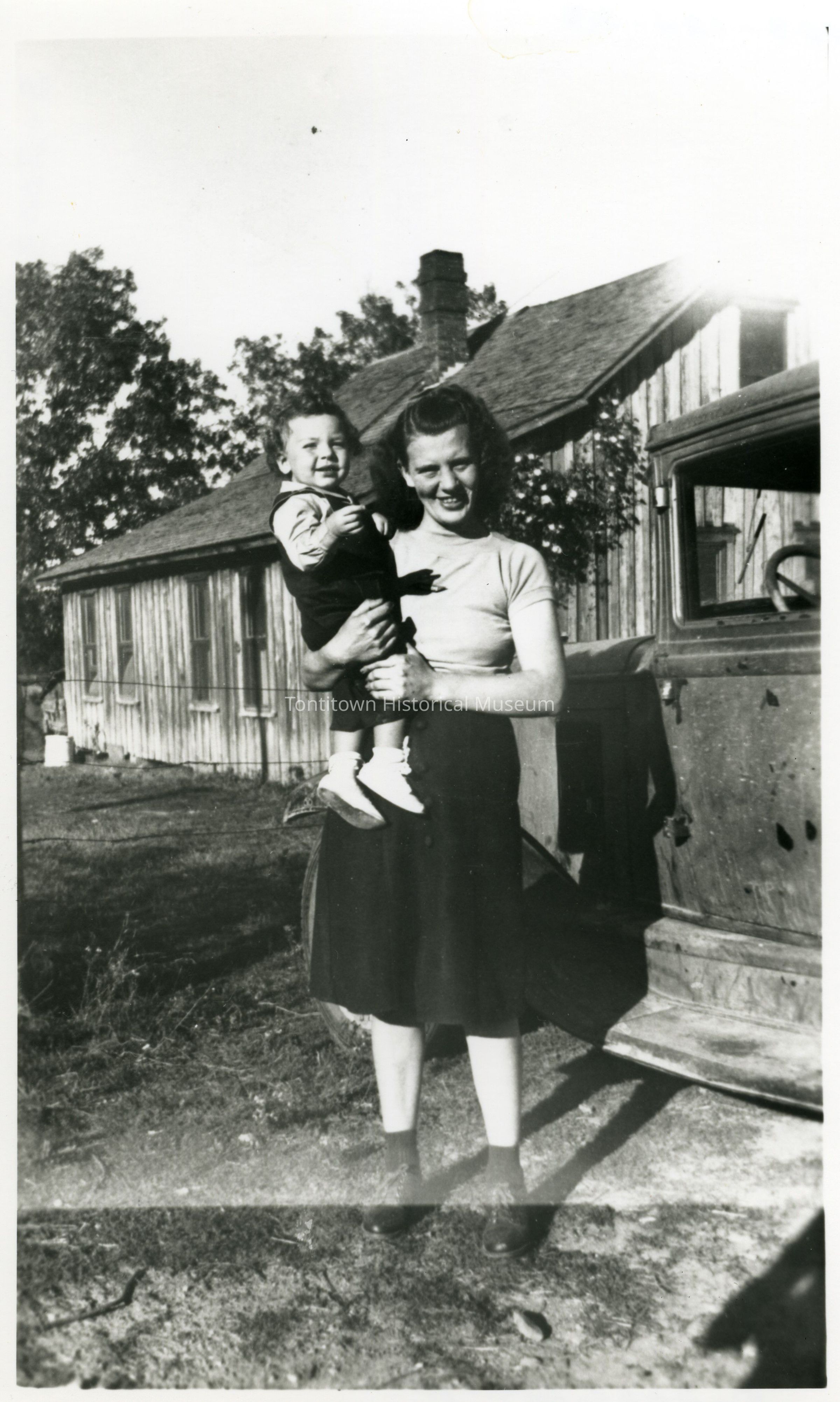 In a black-and-white photograph, a woman stands outdoors, smiling warmly while holding a young child in her arms; they are positioned near an old vehicle, with a rustic wooden house as the backdrop, surrounded by trees under a clear sky, evoking a nostalgic and serene moment in time, possibly from the mid-20th century, capturing a slice of everyday rural life, with the woman wearing a light top and dark skirt, and the child dressed in a small outfit with white shoes, embodying