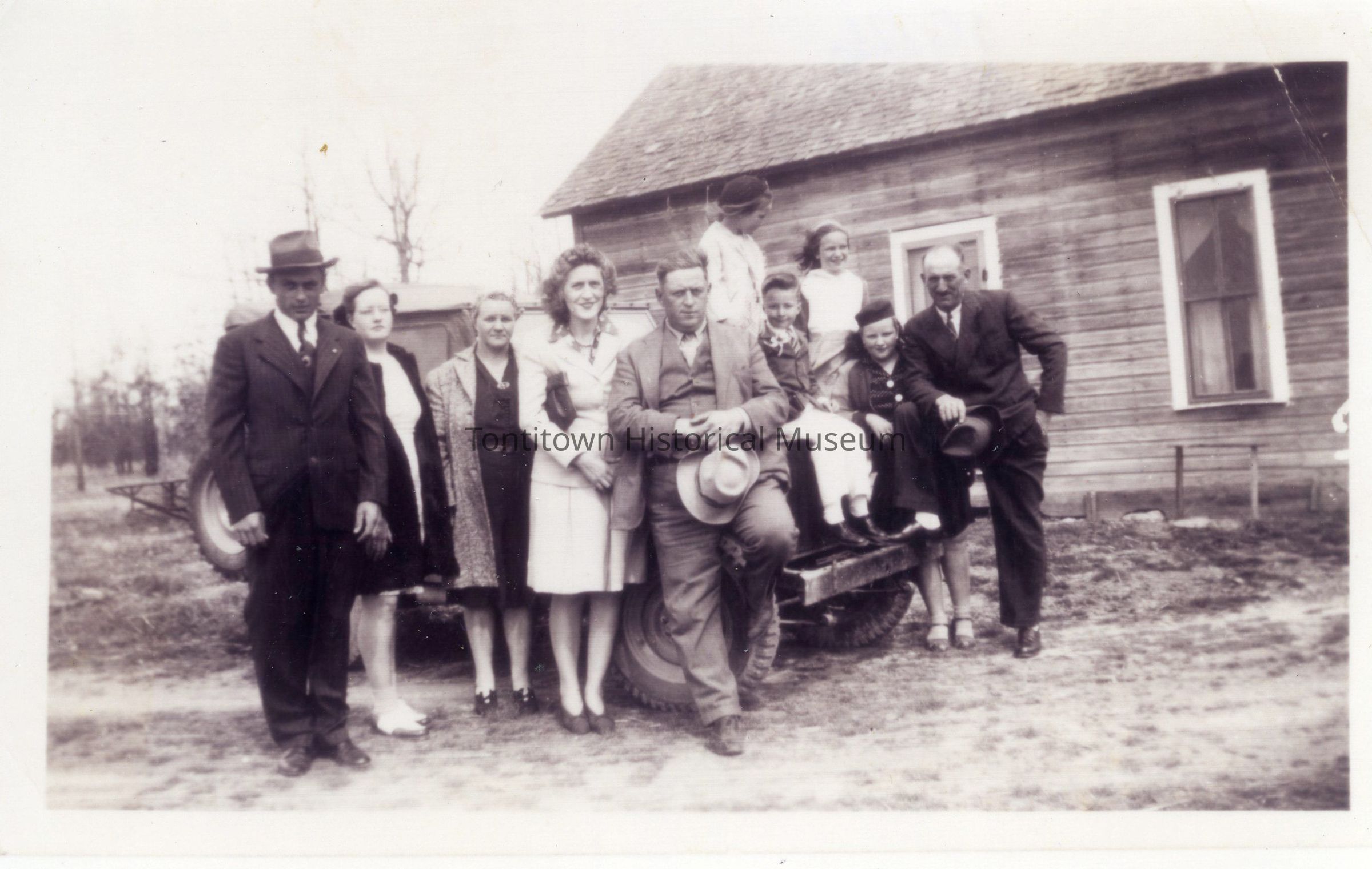 The black and white photo depicts a group of nine individuals, consisting of adults and children, gathered in front of a wooden house, alongside a vintage vehicle. The adults are dressed in formal attire typical of the early to mid-20th century, with coats and hats, while the children appear warmly dressed, some perched on the vehicle's rear. The house in the background is rustic, with a wooden exterior and a simple window, suggesting a rural setting. The scene conveys a sense of family or community