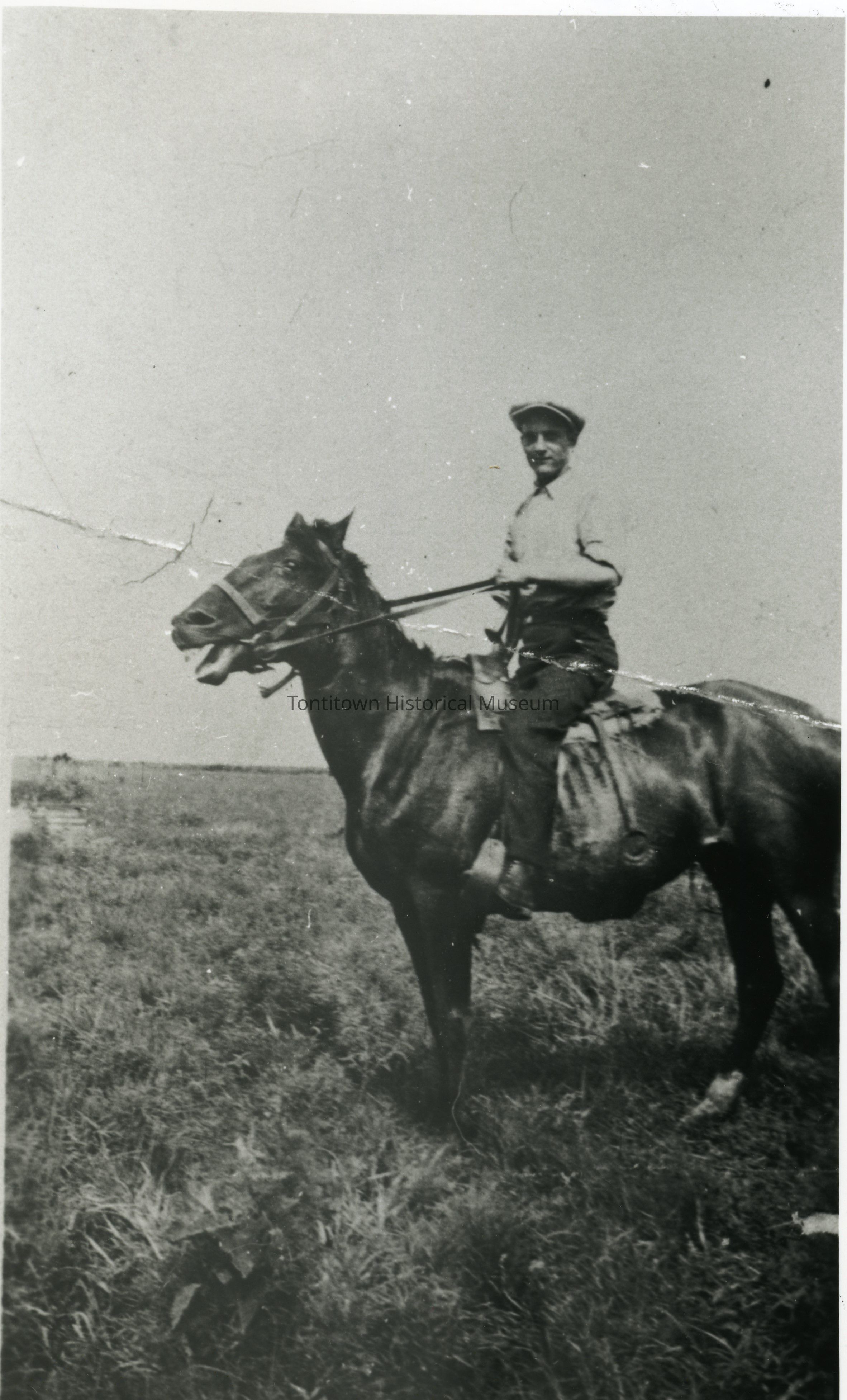 In this vintage black and white photograph, a man is seen confidently sitting in saddle atop a dark open-mouthed horse across a grassy field; man is dressed in a light-colored collared shirt and a cap, as his hands grasp the reins; the horse stands alert and poised, with its head slightly turned; the landscape stretches into the distance under an expansive sky
