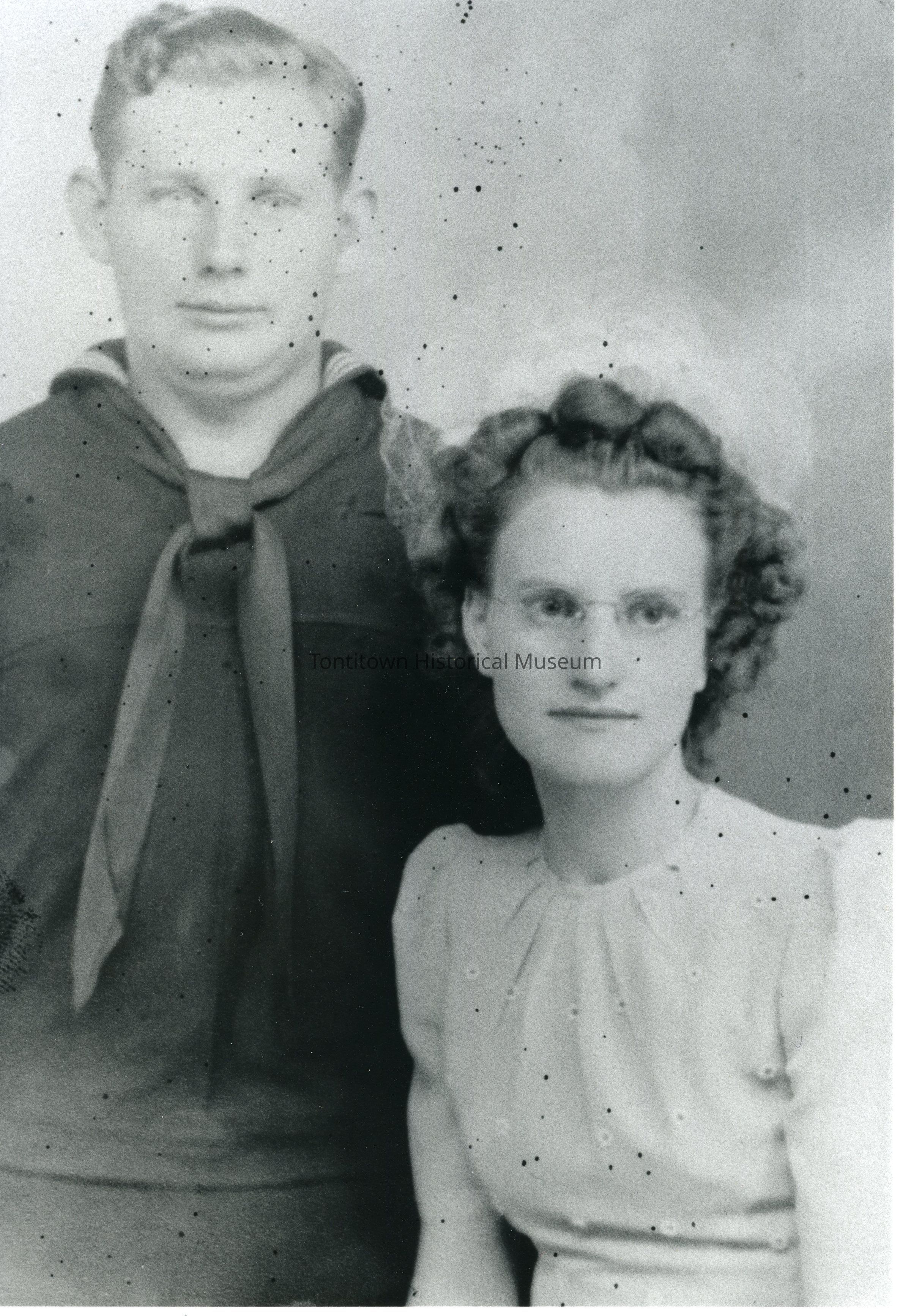 Cigainero-Jaro Wedding: The black and white photograph depicts a man in a naval uniform standing beside a woman dressed in a formal outfit, likely from the mid-20th century, as evidenced by the style of clothing and the photograph's vintage appearance; the man, wearing a dark sailor uniform with a tied neckerchief, while the woman, seated slightly in front of him, wears glasses and a neatly styled hairdo.