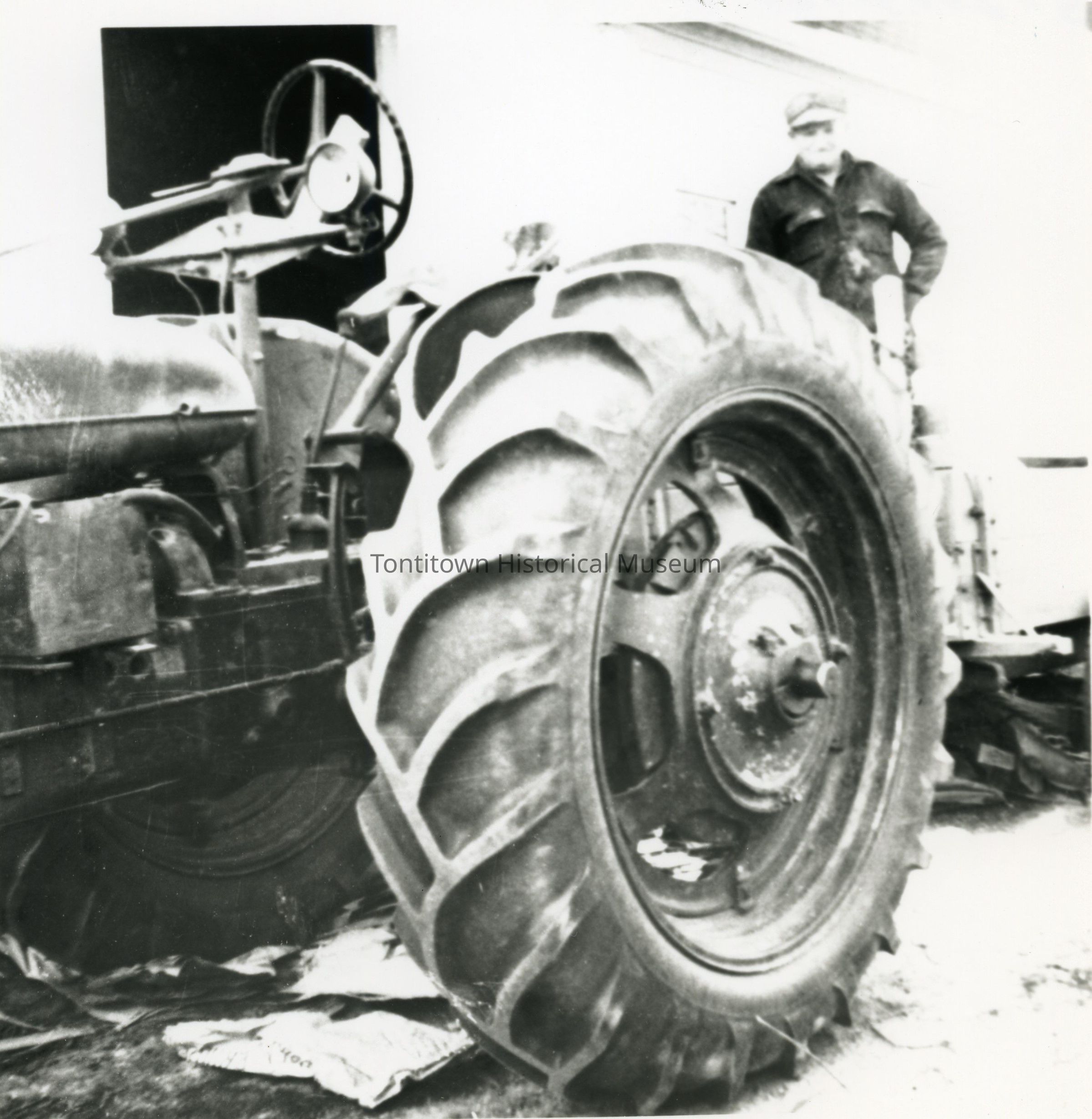 The image shows a large tractor with prominent, textured tires positioned in the foreground, capturing the mechanical details and rugged design typical of agricultural machinery; a man, wearing a cap and dark coverall-type clothing, is standing nearby, possibly overseeing or working with the tractor, that appears to be situated outdoors, perhaps on a farm or rural area, with minimal background elements visible.