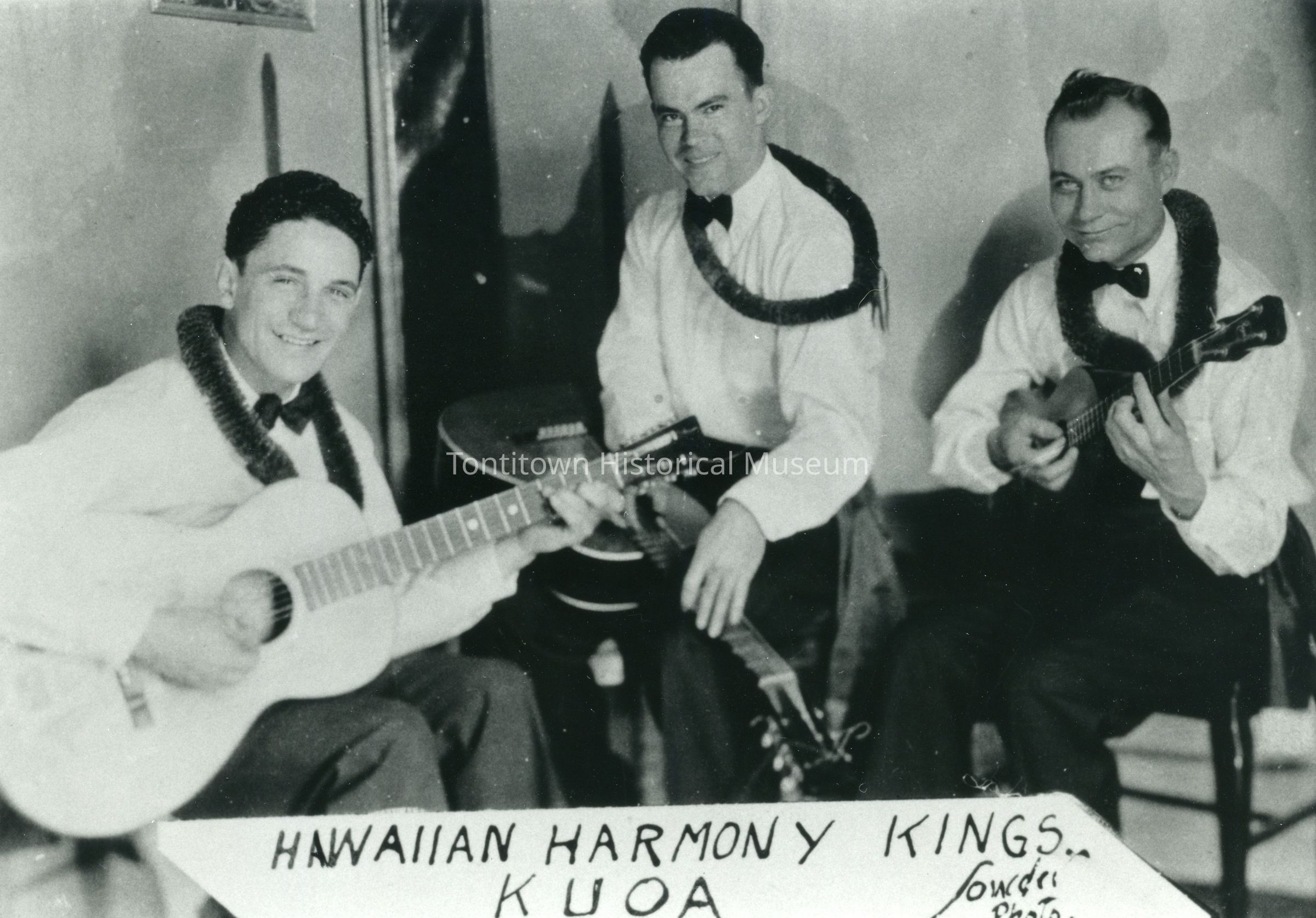 Hawaiian Harmony Kings: In this black-and-white photograph, three musicians from the band "Hawaiian Harmony Kings" are joyously seated with their instruments; the person on the left plays an acoustic guitar while smiling directly at the camera, the middle person manages another string instrument, possibly a fiddle guitar, and the person on the right skillfully strums a ukulele; all three wear leis and are dressed in formal shirts with bow ties, exuding a blend of elegance and casual Hawaiian charm.