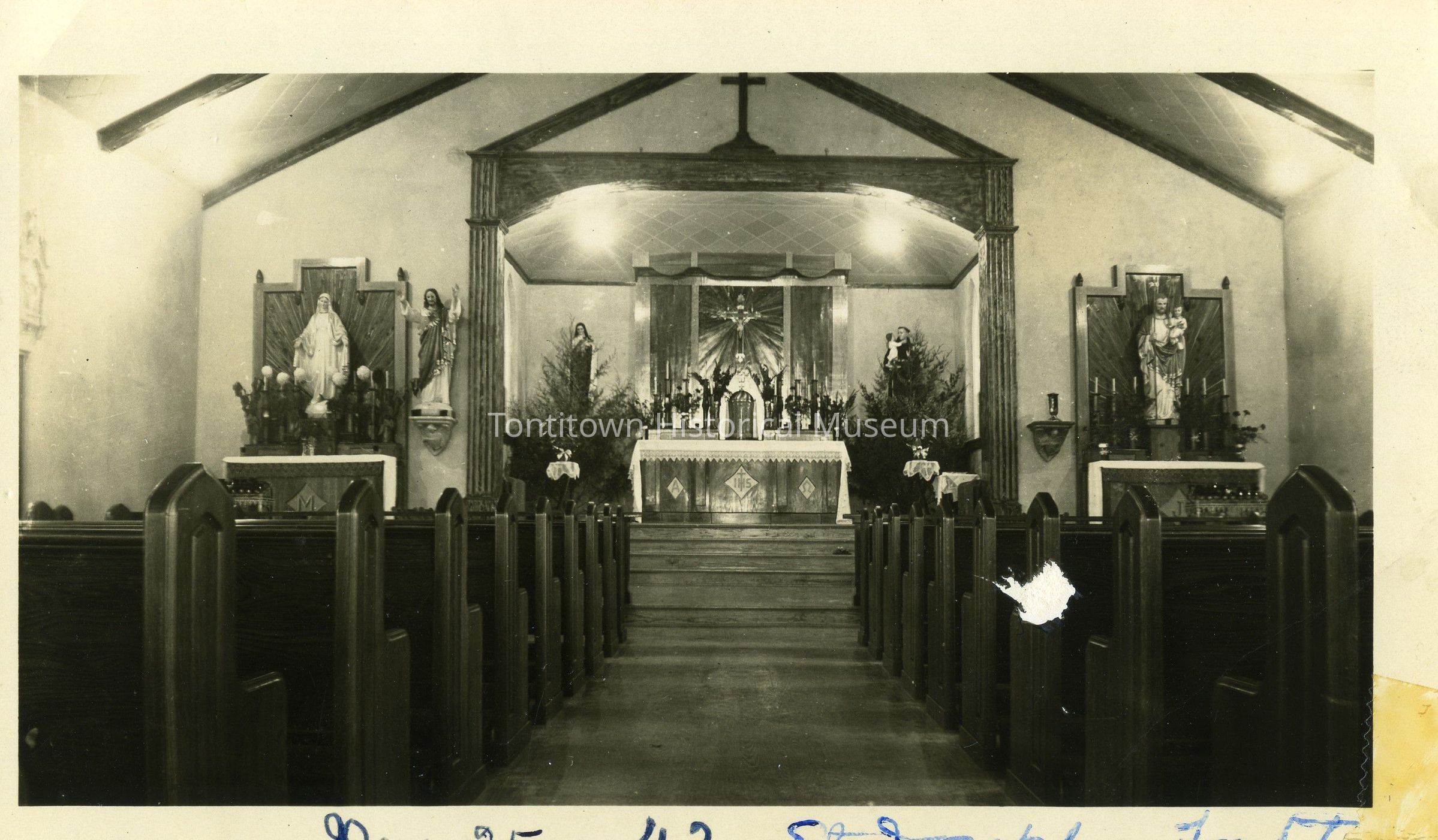 The black-and-white photograph depicts the interior of a church with wooden pews leading up to an ornate altar adorned with religious statues and candles. Two statues flank the altar on each side, and tall, decorated trees add an element of festivity. The altar, elevated and central in the composition, features intricate carvings and a prominent crucifix above it. Light fixtures, hanging from the ceiling, illuminate the space, casting gentle shadows across the wooden surfaces. The architecture is traditional, with a vaulted ceiling