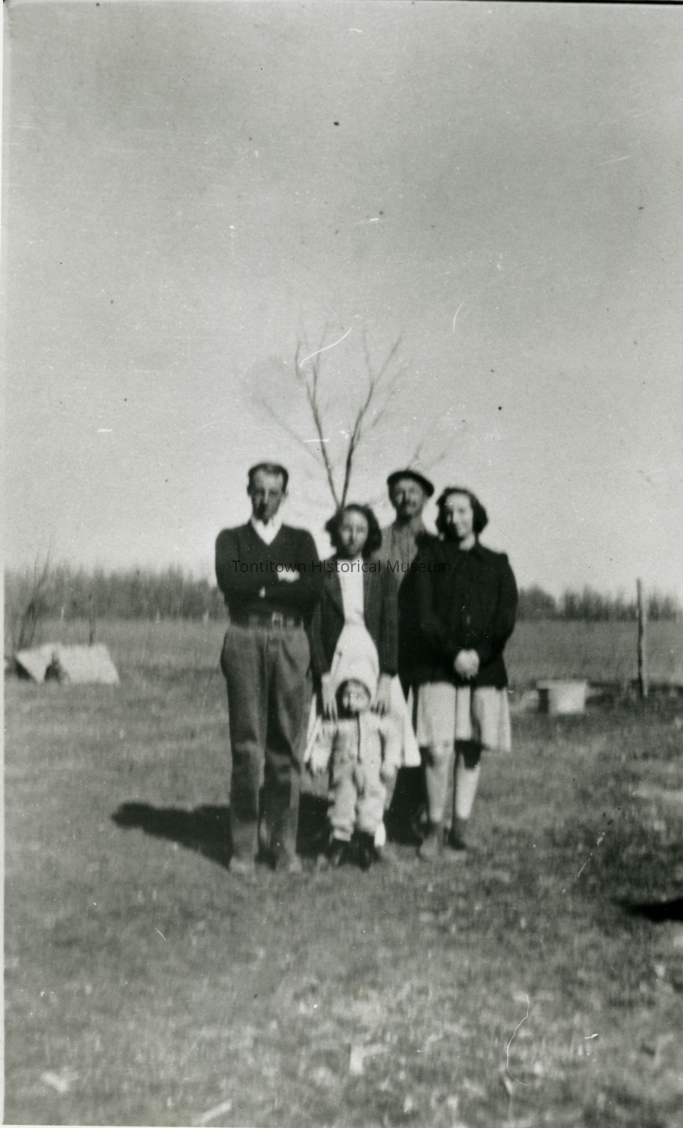 In a black and white photograph depicting a rural setting, four adults and a child are standing outdoors on a patch of open ground, with the adults dressed in typical mid-20th century attire, including sweaters, coats, and hats; the child, positioned in front of the group, wears a cap and warm clothing suitable for cool weather. Behind them, a bare, leafless tree stands against a backdrop of blurred, expansive landscape, while other distant trees or foliage create a natural horizon line, adding