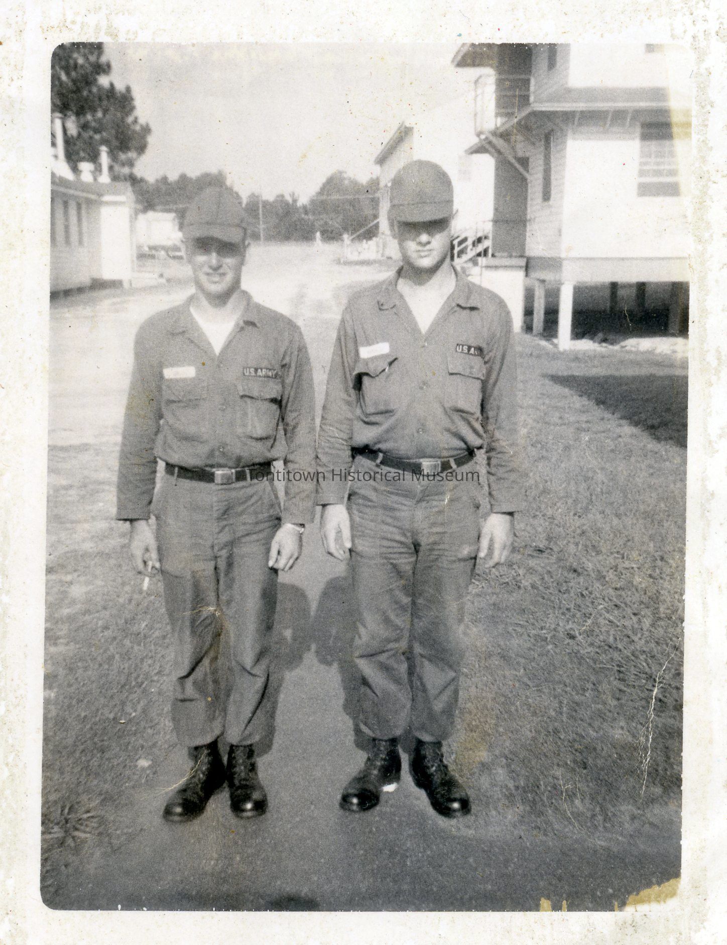 The black and white photograph features two men standing side by side, both dressed in matching military uniforms, with caps and boots, labeled "U.S. Army." They are positioned on a paved path, within a military base, as shown by barrack-style buildings in the background. Their expressions are neutral, and they appear to be posing formally for the photograph. Man on left holds cigarette in his right hand. Anthony Ranalli and Kenny Green.