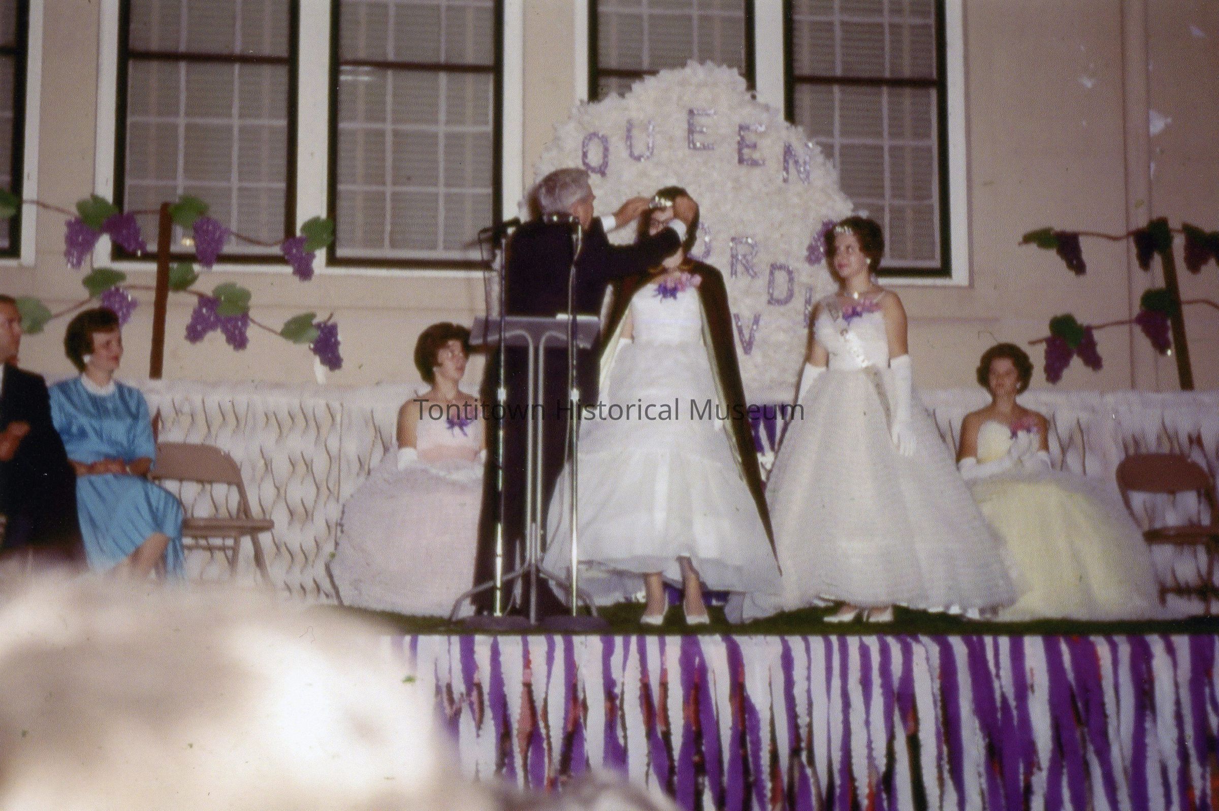 The image depicts a formal ceremony on a decorated stage where an individual is crowning a young woman in a white gown with a cape, while four other women in elegant dresses and a couple seated on chairs observe the event; the background is adorned with grape-themed decorations and the words "Queen" and "Court" can be seen, suggesting a pageant or festival setting.