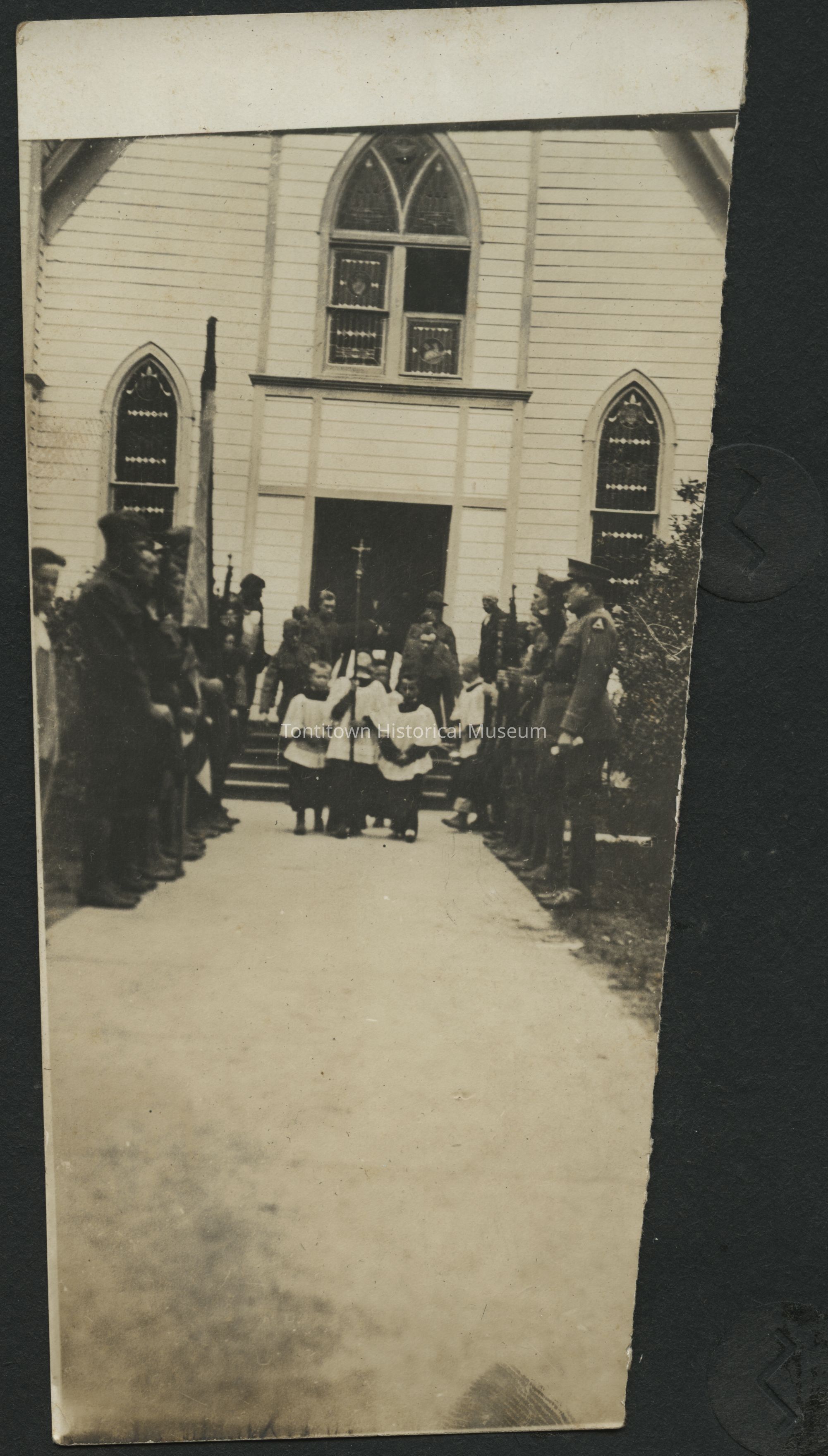 In a sepia-toned photograph, a group of young altar servers stand in front of a church, dressed in traditional robes, surrounded by uniformed soldiers lining the pathway, possibly forming a guard of honor; the church features distinct Gothic-style windows, and a cross is visible, suggesting a religious ceremony or procession as people gather in observance, creating a historical atmosphere indicative of a significant communal or church-related event, captured in a moment that reflects a blend of solemnity and tradition.