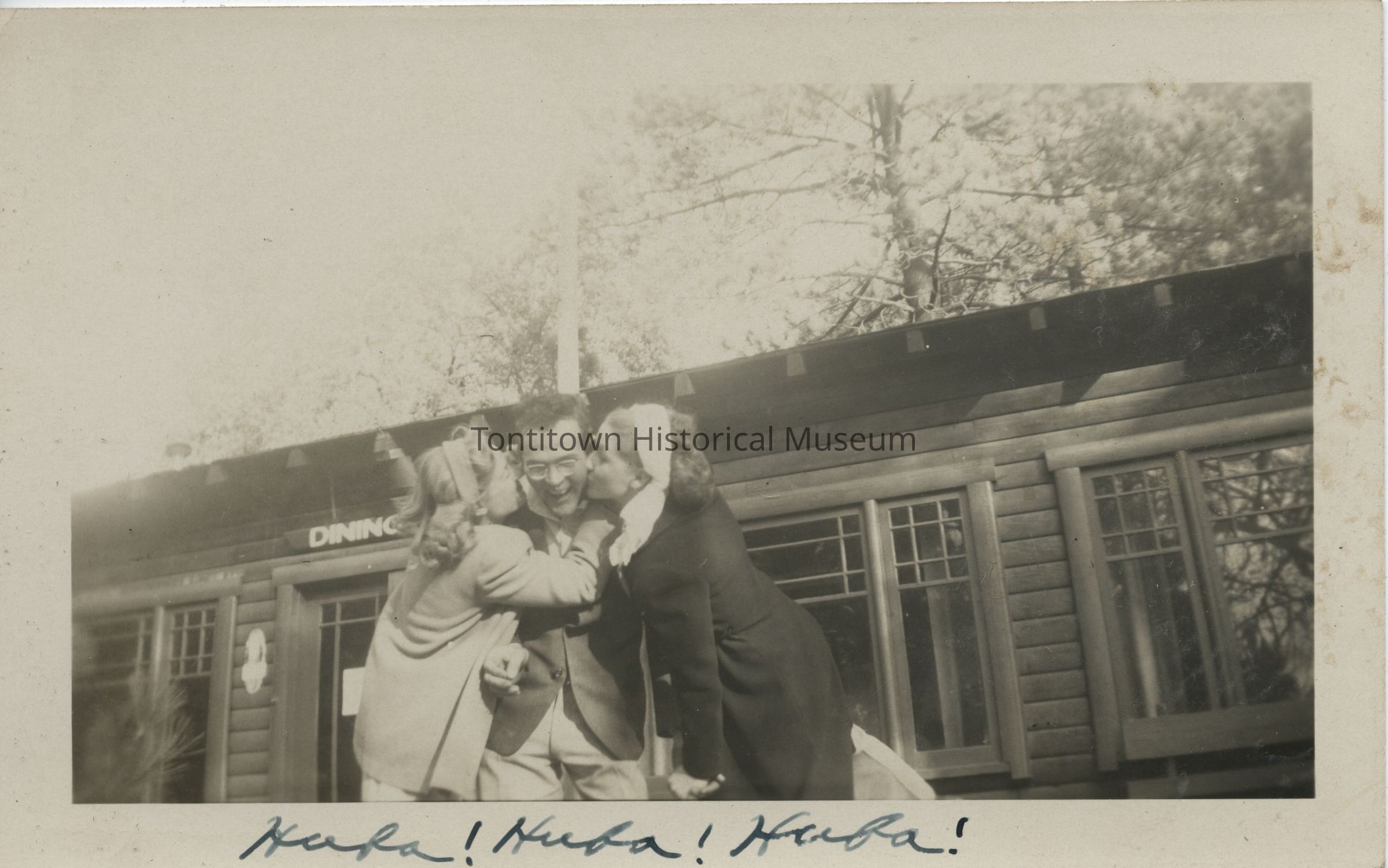 In this charming vintage photograph, three people are engaged in a joyful moment outside a rustic log cabin, with the two individuals on either side planting affectionate kisses on the cheeks of the delighted person in the middle; the background includes trees and the partial signage of the cabin, which reads "DINING," adding a quaint and nostalgic feel to the scene, while the handwritten words "Hubba! Hubba!
