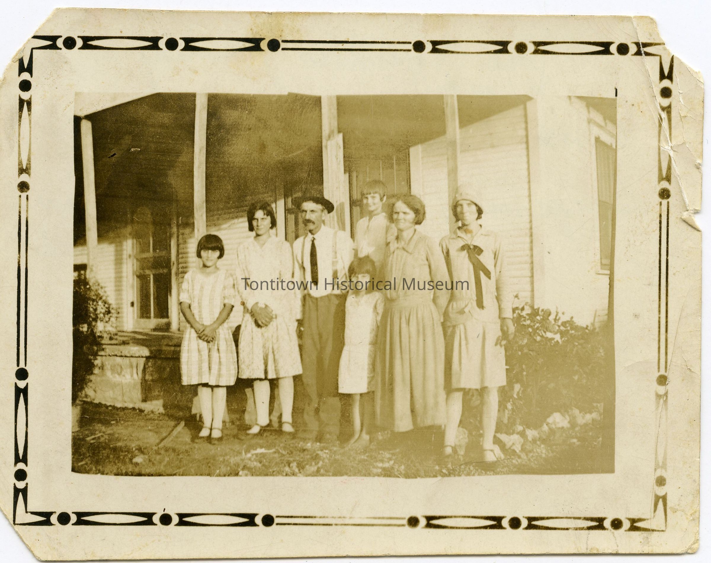The image depicts a vintage photograph of a family group, consisting of seven individuals, including adults and children, standing closely together in front of a porch attached to a wooden house with visible siding and a window, surrounded by a rustic environment that evokes a sense of history and tradition. The adults are dressed in period-appropriate clothing, with the men in hats and suspenders, and the women in dresses, some with hats, while the children are similarly attired, suggesting a formal occasion or family gathering.