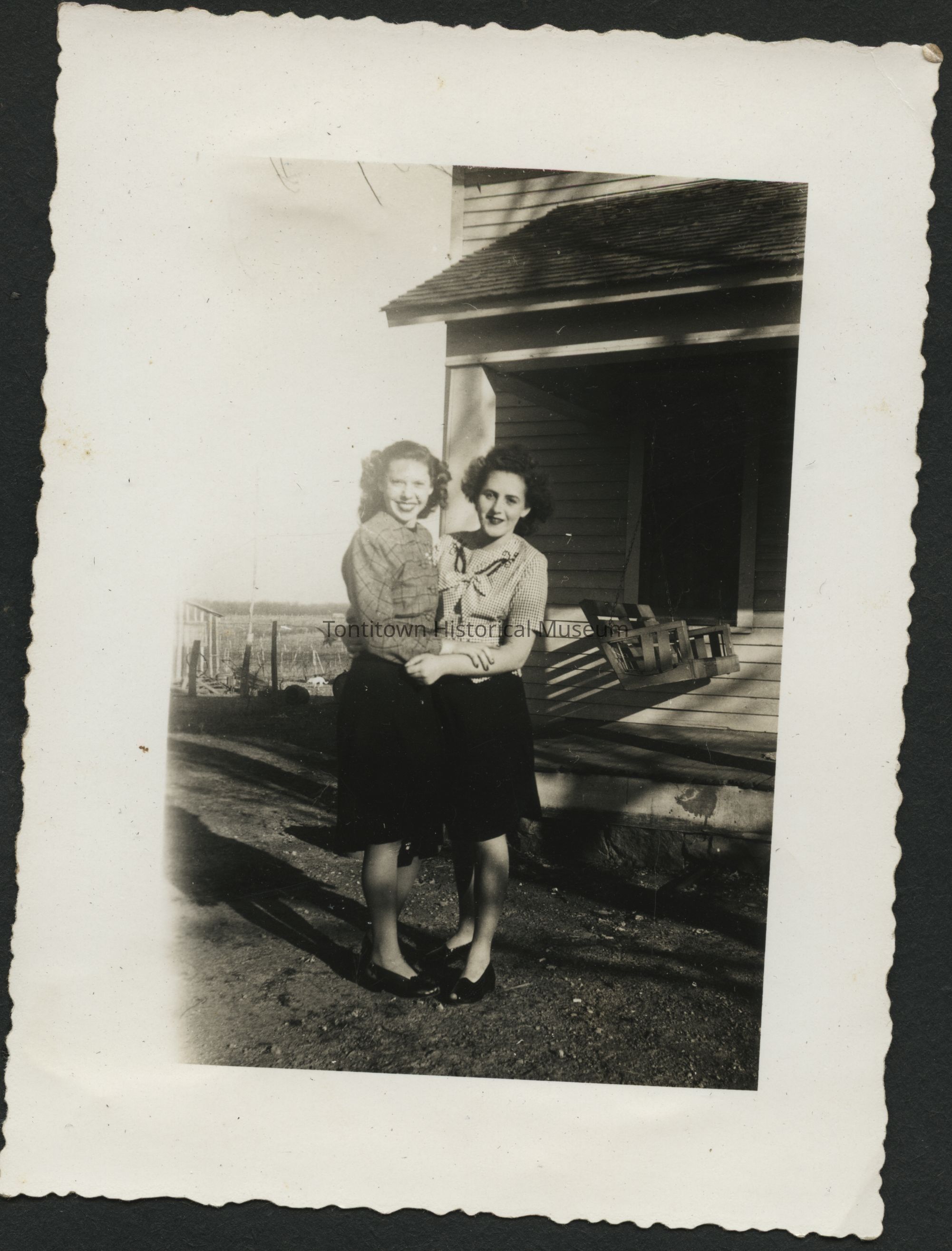 In this vintage black-and-white photograph, two women stand close together, smiling warmly as they embrace each other in front of a wooden house with a porch swing visible in the background. Both are dressed in knee-length skirts and blouses, embodying a mid-20th-century fashion style. The sunlight casts gentle shadows on the ground, suggesting the photo was taken outdoors on a clear, sunny day.