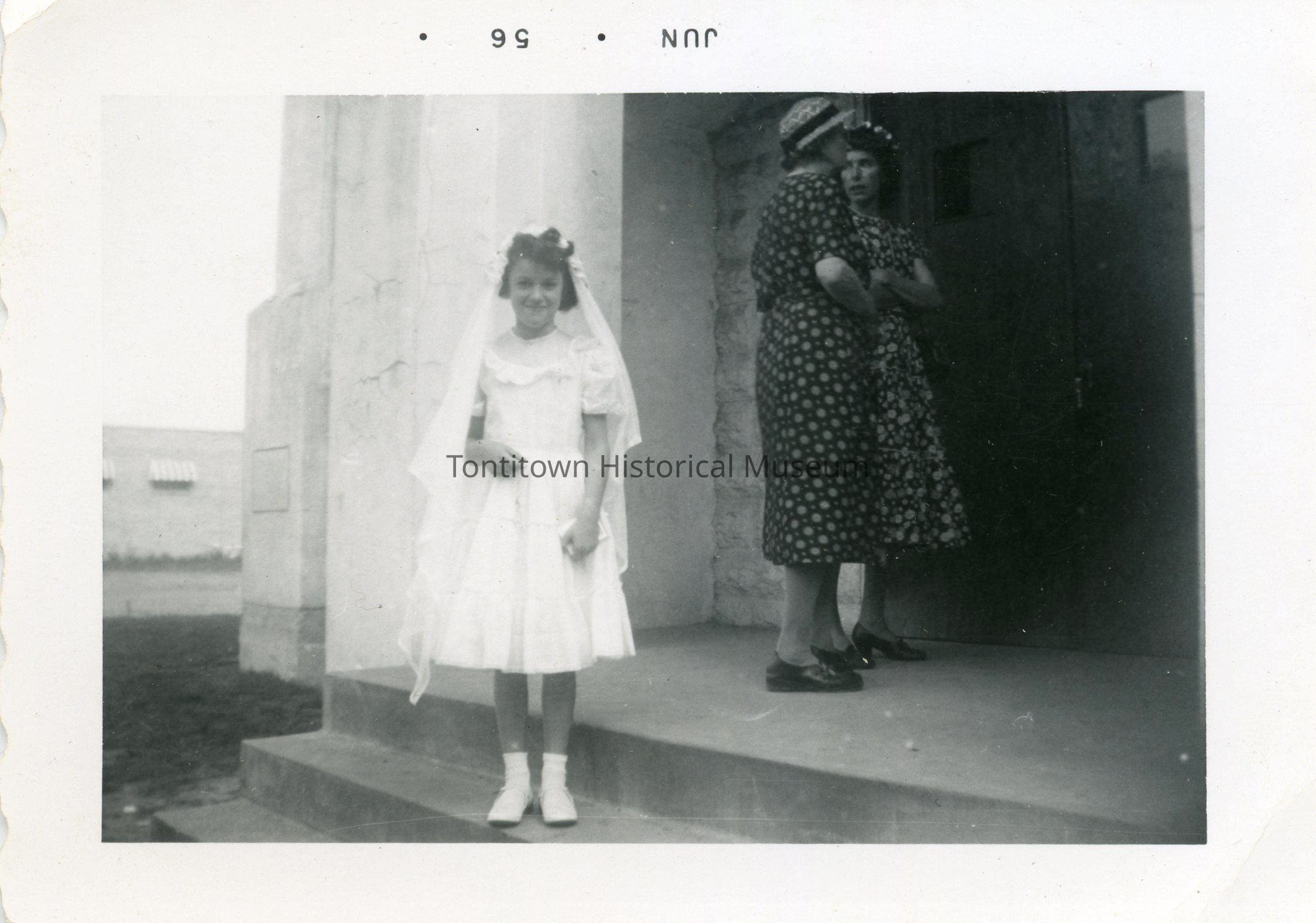 In a vintage photograph marked June '56, a young girl Agatha Penzo, stands on the steps of a building, dressed in a white ceremonial dress with a veil, prepared for a first communion. She looks directly at the camera. To the right, two women engage in conversation, dressed in patterned dresses typical of the era, while standing by the entrance.