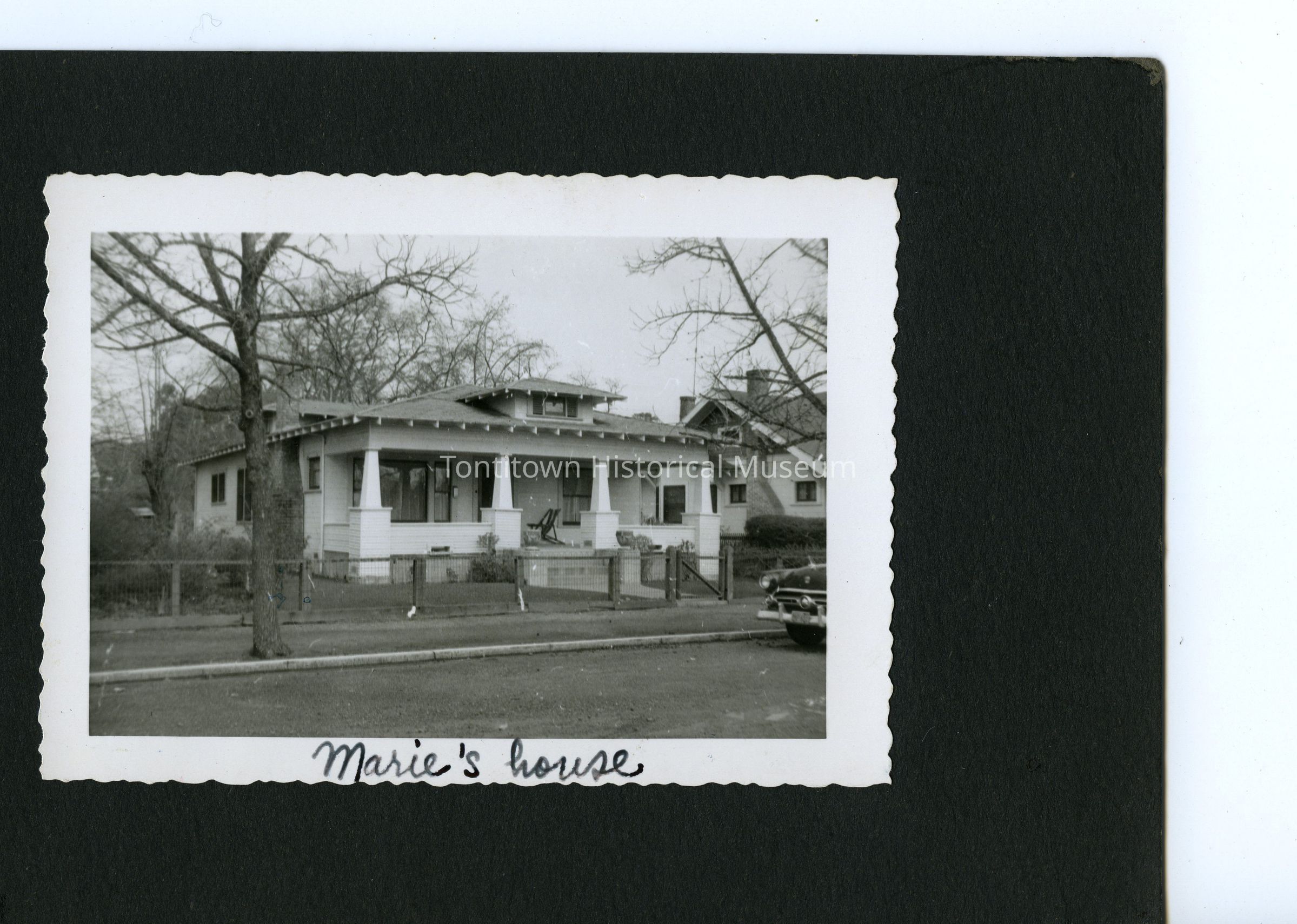 The black-and-white photograph showcases a quaint, single-story house with a covered porch supported by sturdy columns, surrounded by leafless trees and partially enclosed by a low fence, suggesting it might be late autumn or early winter; a vintage car is parked on the street to the right side of the house, while the handwritten text, "Marie's house," is inscribed on the bottom border.