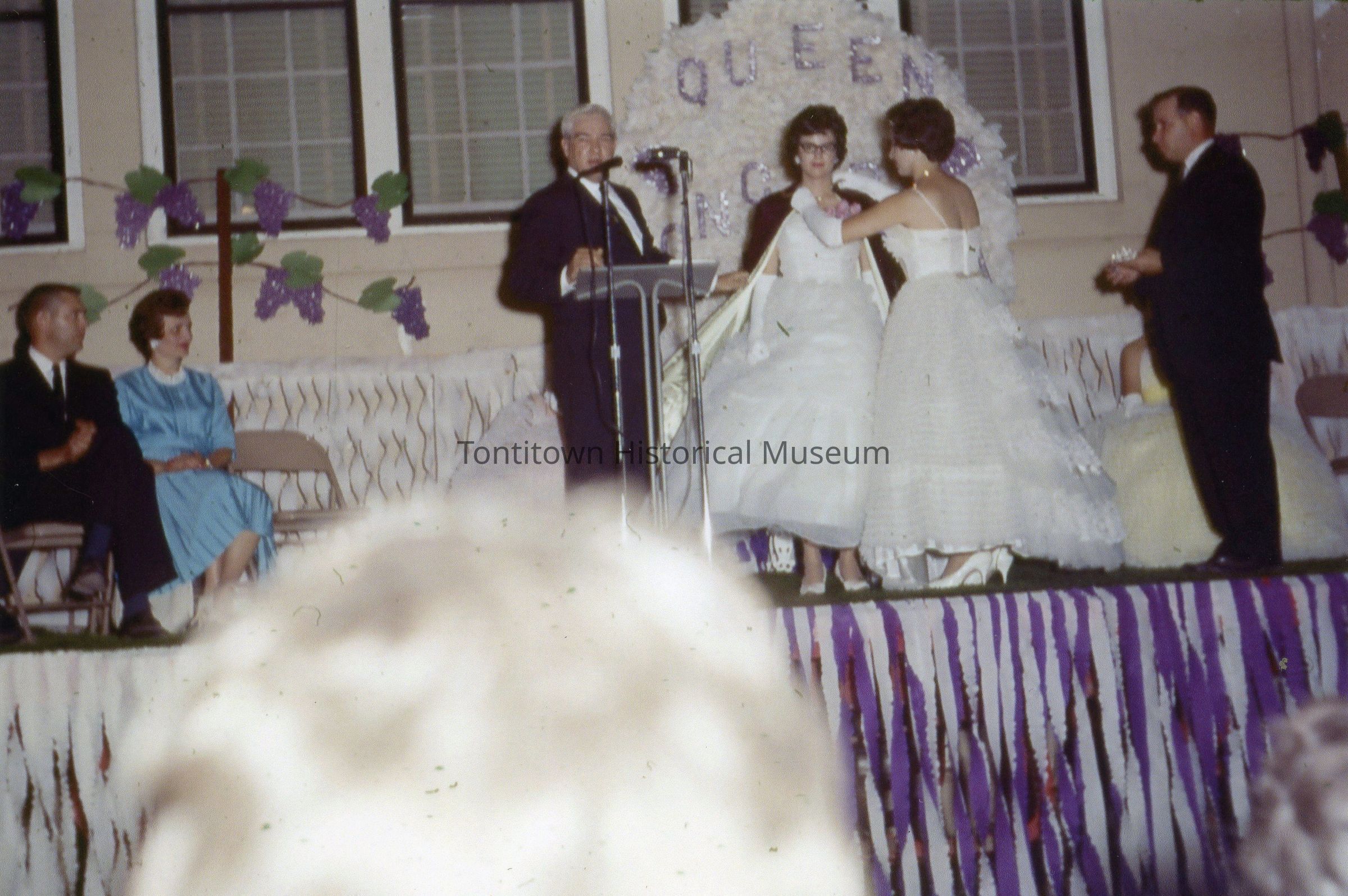 The image depicts a vintage scene of a pageant or ceremonial event in which a woman in a white gown and gloves is crowning another woman, also in a white gown, while a man at a podium, likely the host or announcer, speaks into a microphone; the background is decorated with a large sign featuring the word "QUEEN" and grapevine motifs, suggesting a theme or festival, and several people are seated, observing the event, including a woman in a blue dress and a man