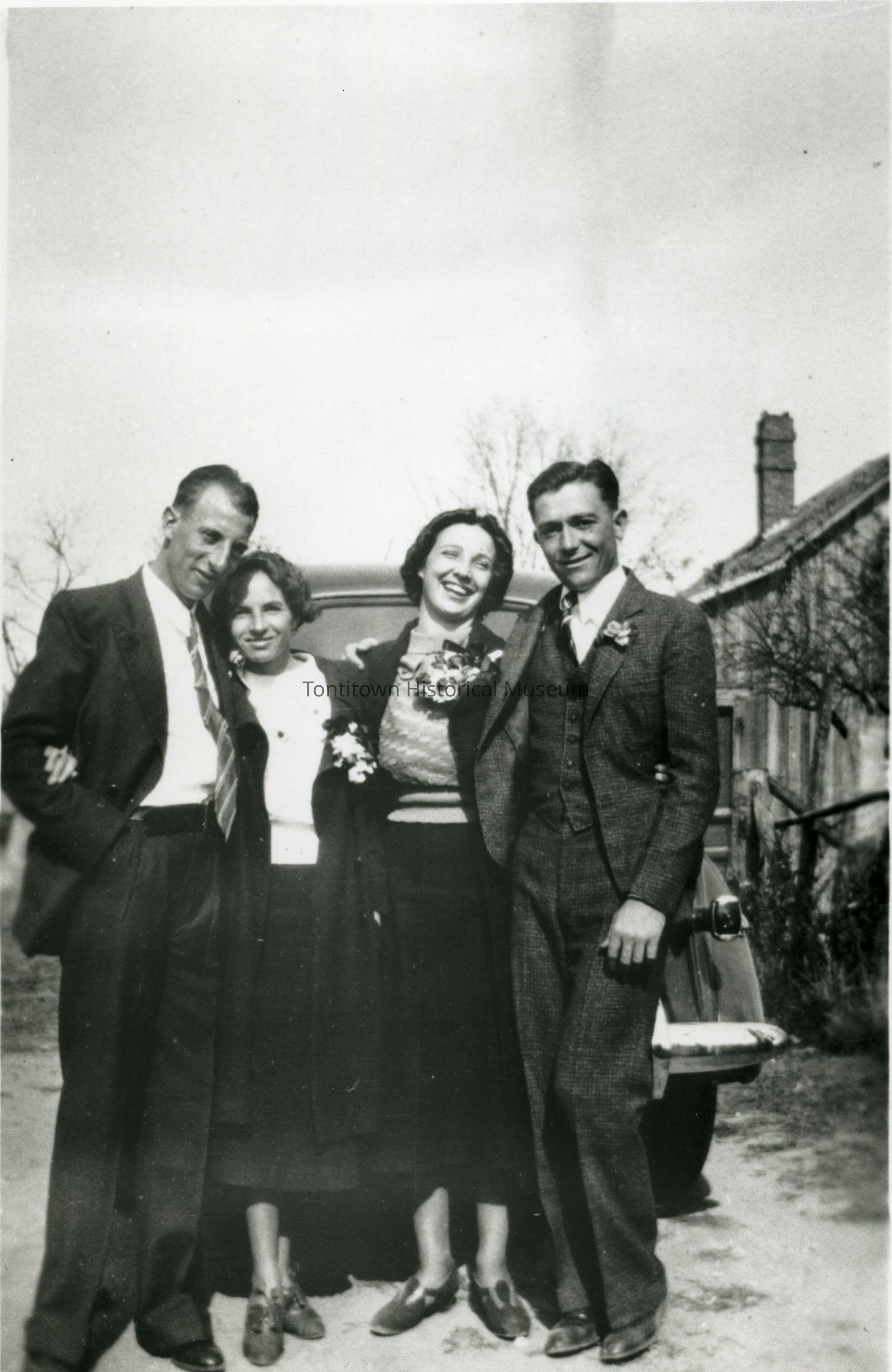 In this vintage black and white photograph, two men and two women stand closely together with their arms linked, displaying joyful expressions as they pose in front of an automobile on what appears to be a country road, while the men don suits with ties and the women wear elegant coats, adorned with floral corsages, suggesting a formal or festive occasion; in the background, sparse trees and a building with a chimney are visible against a partly cloudy sky, adding a rustic charm to the scene captured in an era when