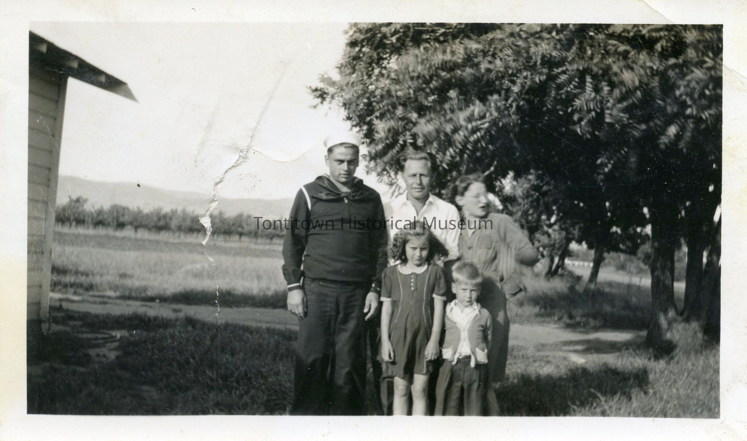 Black-and-white photograph features an outdoor portrait with five individuals between a tree and a home: two men, one dressed in what appears to be a sailor uniform, standing on the left, while another man in light clothing stands beside him; slightly in front of them, a young girl in a dark dress with a white collar, and a smaller boy with a light shirt and dark pants are positioned; a woman stands close to the group, gesturing animatedly, all framed by a background of trees and a distant landscape.