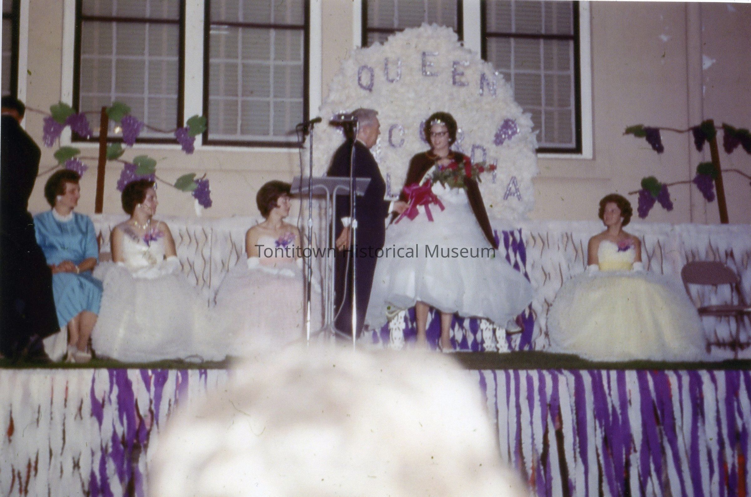 The image depicts a formal event likely celebrating a beauty or cultural pageant, where a woman stands in the center, wearing a white dress and holding a bouquet, while receiving recognition on stage; she is flanked by four seated women, each dressed in elegant gowns, with purple floral decorations and a backdrop displaying the word “QUEEN”; other event details, such as a podium with microphones and festival-like decorations, suggest a celebratory, formal occasion with queenship or similar titles being conferred, indicating