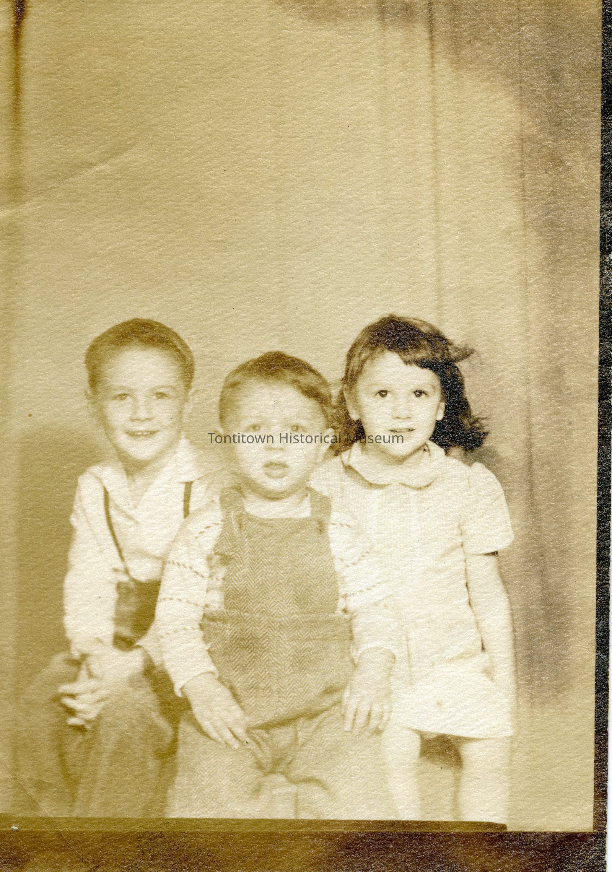 The sepia-toned vintage photograph features three young children posed side by side; on the left, a boy wearing suspenders and a collared shirt sits with a warm smile, while in the center, a toddler in overalls and a patterned sweater gazes directly at the camera with a curious expression; on the right, a girl in a light-colored dress with a Peter Pan collar, her hair gently tousled, poses with a composed and thoughtful look, capturing a moment of nostalgia and familial warmth