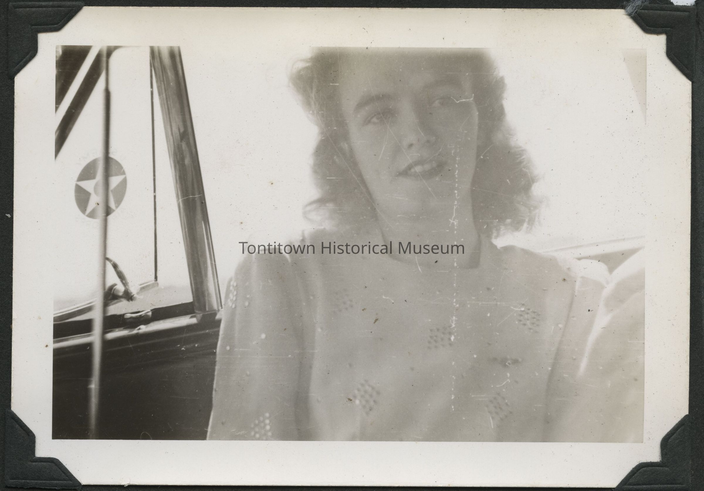 In this vintage black and white photograph, a young woman with curly hair is depicted sitting in what appears to be a vehicle, perhaps a car, wearing a dress adorned with a subtle pattern, as sunlight filters into the interior, creating a soft and nostalgic ambiance; the window behind her shows a circular decal featuring a star, suggestive of a specific location or establishment that offered a backdrop during this captured moment in time.
