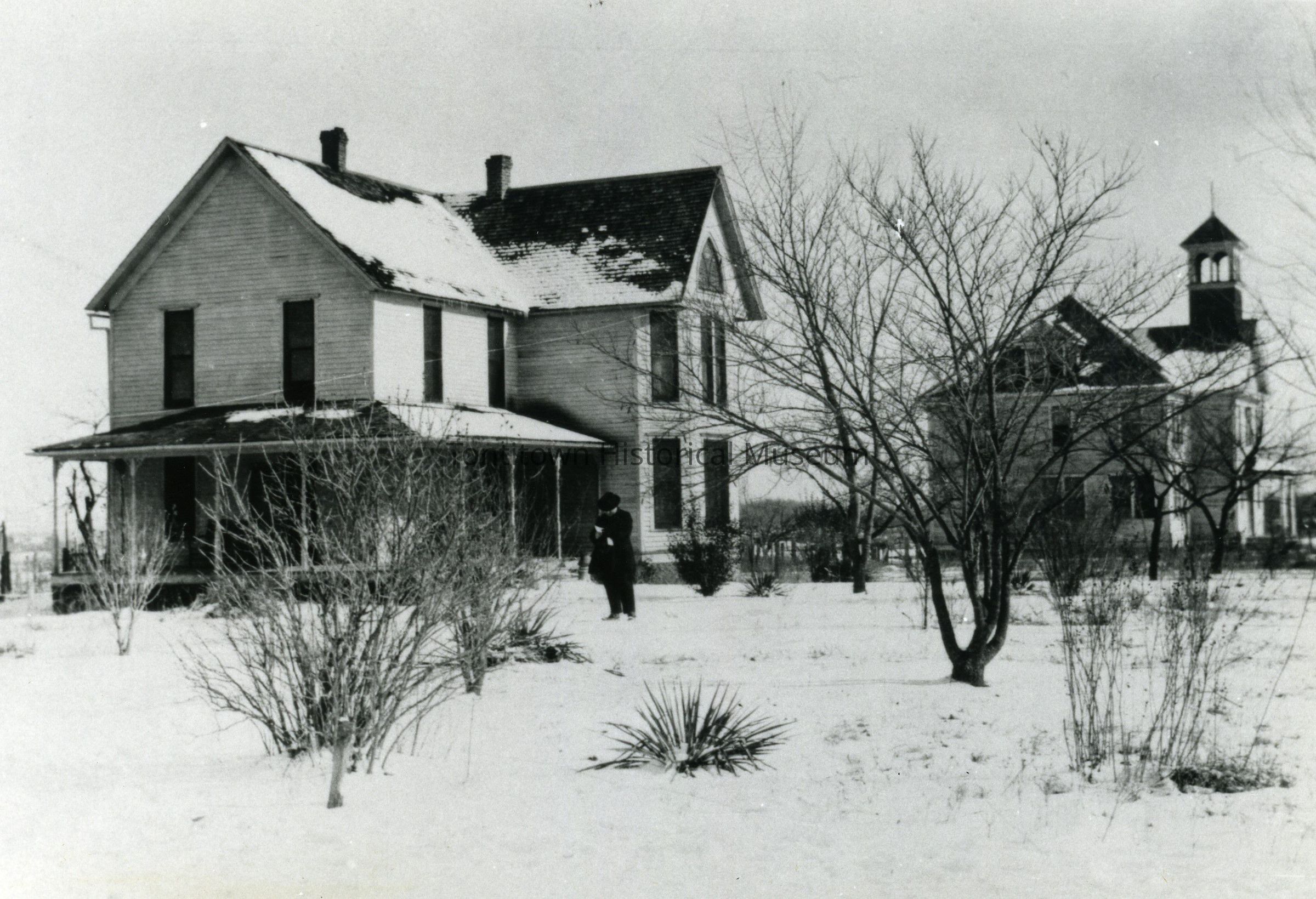 The black and white photograph depicts a snow-covered landscape featuring a two-story wooden house with a covered porch, flanked by bare trees and bushes in a winter setting; a solitary figure stands in the foreground, wearing dark clothing, adding a sense of scale and perspective to the scene; in the background, another structure with a steeple is visible, hinting at a rural or small-town setting; the contrast between the dark silhouettes of the buildings and trees against the bright snow creates a stark and nostalgic