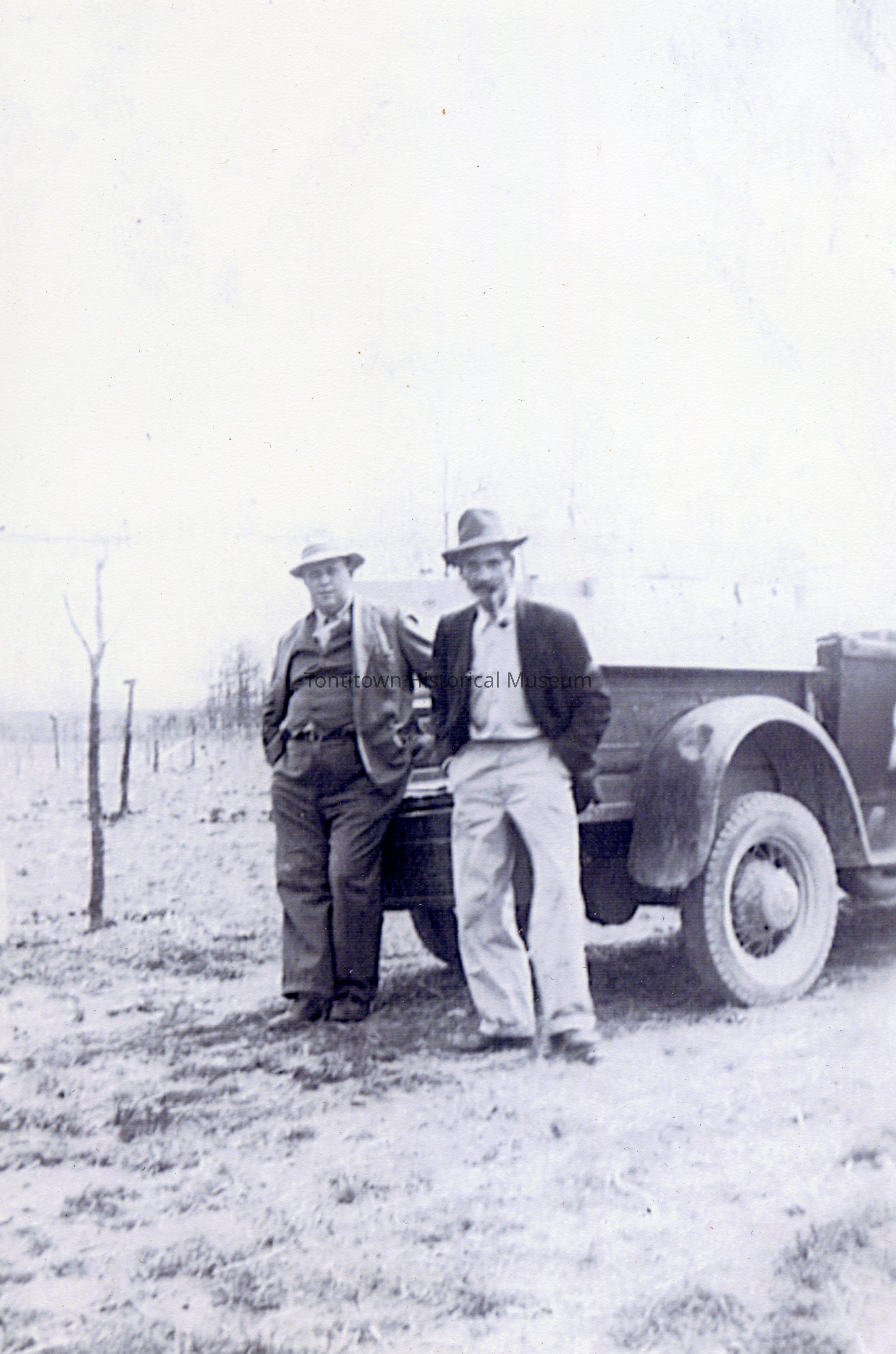 In this vintage photograph, two men are seen standing confidently next to an old car in an open, barren landscape; each man is casually dressed in a suit, one in a three-piece with a vest and the other in a more relaxed style, both with hats that cast a shadow over their faces, giving an impression of the fashion typical of a bygone era, while the sepia tone and muted background evoke nostalgia, hinting at stories of rural life and travel during an earlier time, possibly reflecting
