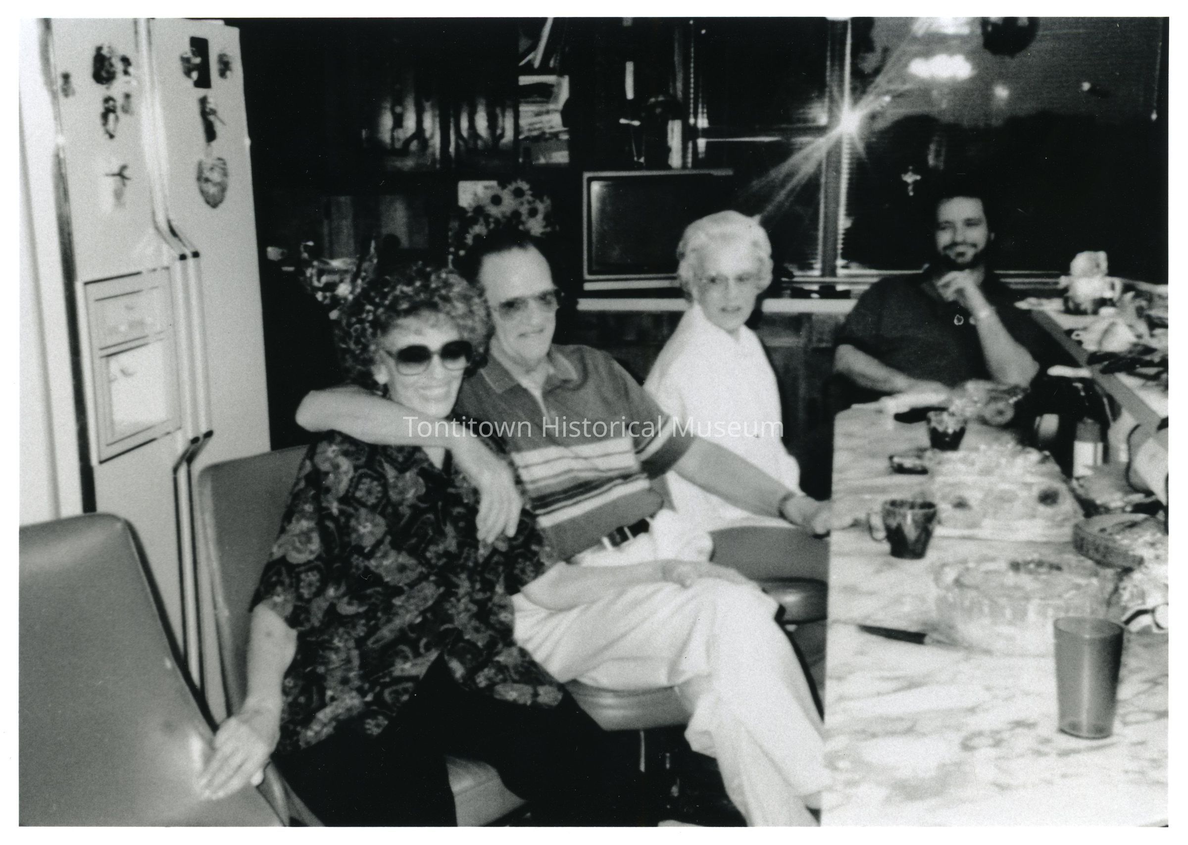 Family: In this black-and-white photograph, four people are gathered around a dining table in a cozy kitchen setting, where a woman in sunglasses, patterned blouse, and curly hair affectionately wraps her arm around the man beside her, who is wearing a striped shirt and glasses; seated next to them is another older woman in a white blouse who looks toward the camera with a pleasant expression, while a man in the background, seated at the table with a beard and dark shirt, has his hand thoughtfully resting against his chin.