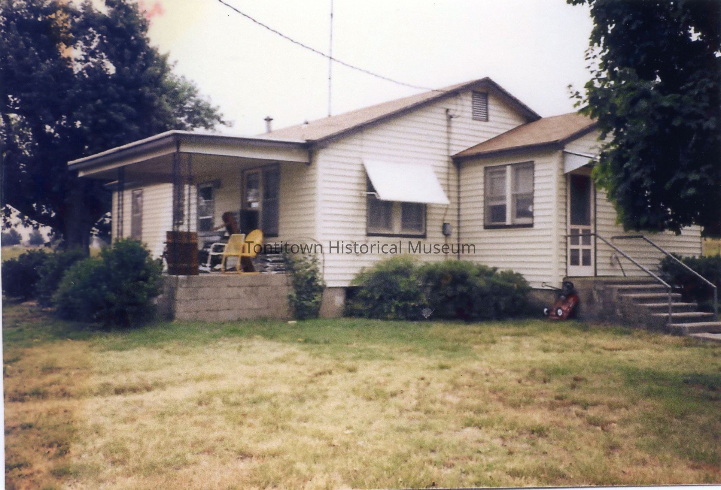 House: The image depicts a modest, single-story house with light-colored siding and a sloped roof, featuring a covered porch adorned with a couple of chairs, resting atop a brick foundation; lush, green bushes line the front of the home, providing a gentle contrast to the neatly manicured lawn that extends into the foreground.
