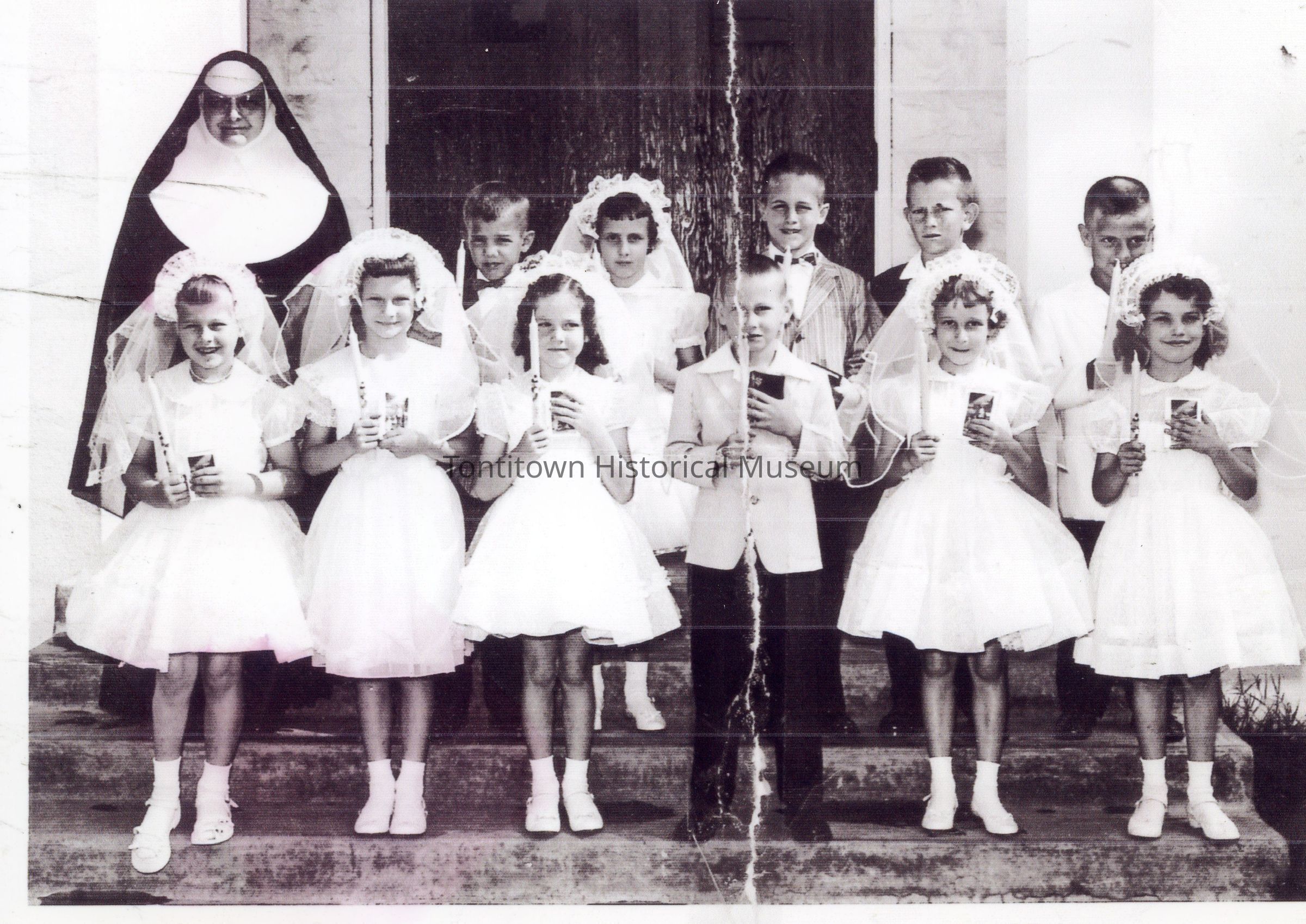 Communion: In the black and white image, a group of young children dressed in formal attire, some wearing white dresses with veils and others in suits, pose on a set of stairs for a photograph alongside a nun clad in a traditional habit, creating a serene and solemn atmosphere that suggests a special religious occasion or ceremony, such as a First Communion.