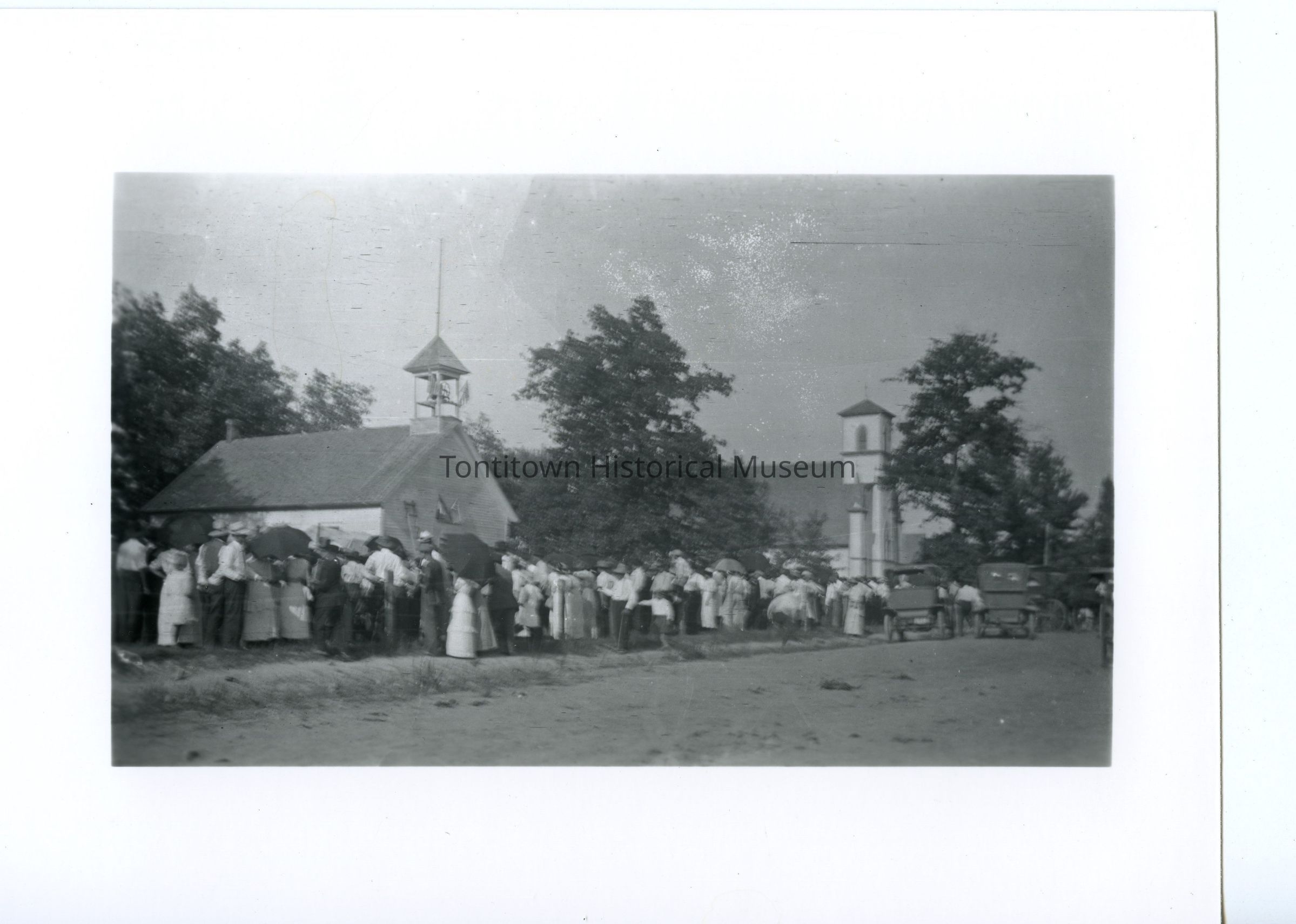 The image depicts a historical scene with a large group of people gathered outside a small church with a bell tower, possibly for a community event or religious service. The attendees, dressed in early 20th-century attire, are standing along the dirt road holding umbrellas, suggesting sunny weather conditions. In the background, another larger building with a tower is visible among the trees, and several vintage automobiles are parked along the road. The overall atmosphere reflects a communal gathering, capturing a moment from a bygone era with