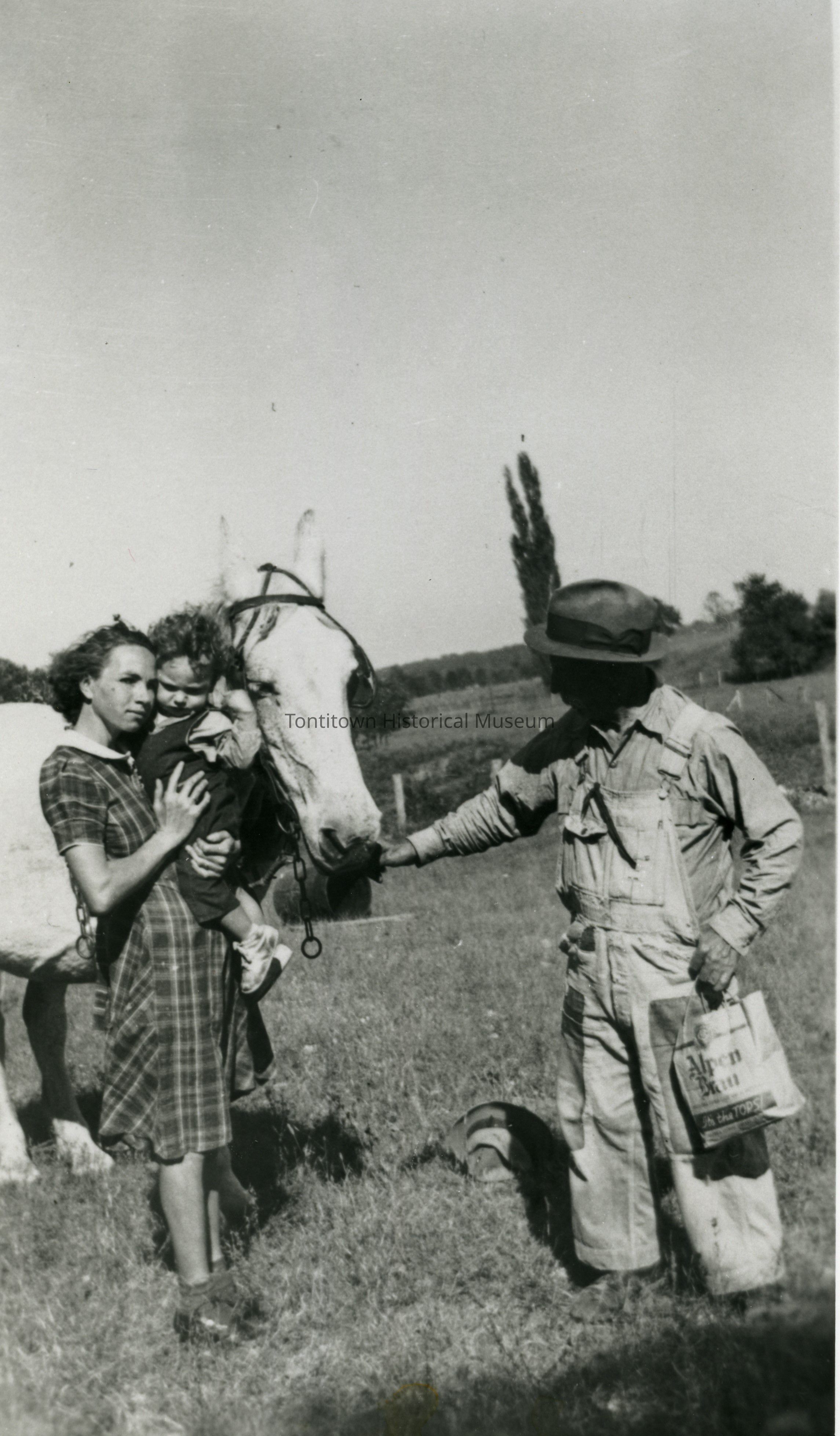 In the black and white photograph, a woman in a checkered dress stands on a grassy field with a serene expression, holding a young child close to a white horse, which they are gently petting. On the right side of the image, a man dressed in work clothes and a hat, carrying a bag that reads "ABERN," stretches his hand toward the horse, creating a peaceful scene of interaction in a rural setting. The background features a few trees and a wide expanse of sky,