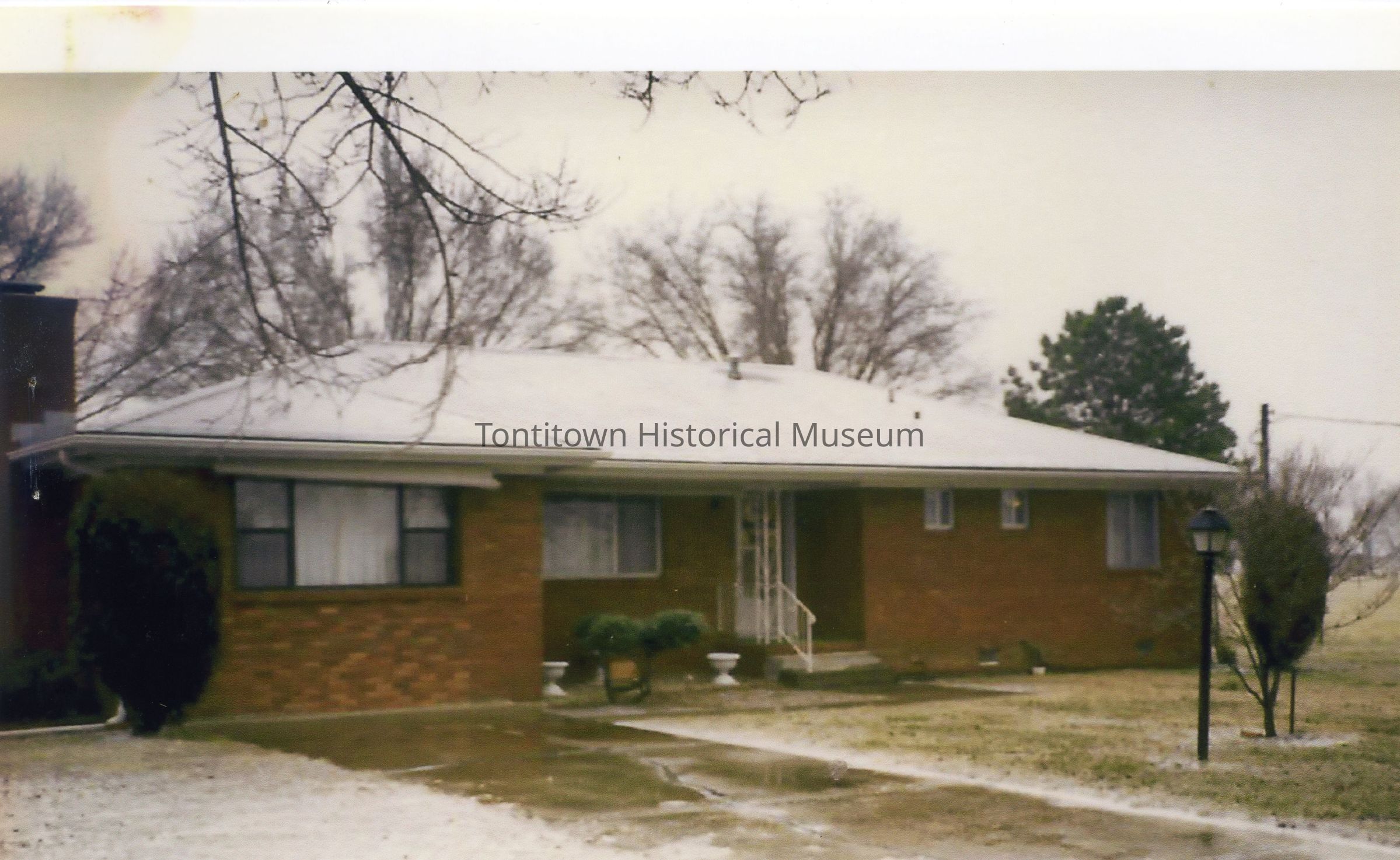 House: In the image, a modest, single-story brick house is depicted with a white, snow-dusted roof, featuring a small porch with decorative railing and two large windows on either side, each covered with sheer white curtains, while a narrow concrete path leads through the sparse, lightly frosted front yard, bordered by a pair of small, manicured bushes and bare-limbed trees that frame the scene, suggesting a cold, wintery atmosphere; additionally, a traditional street lamp stands to the right,