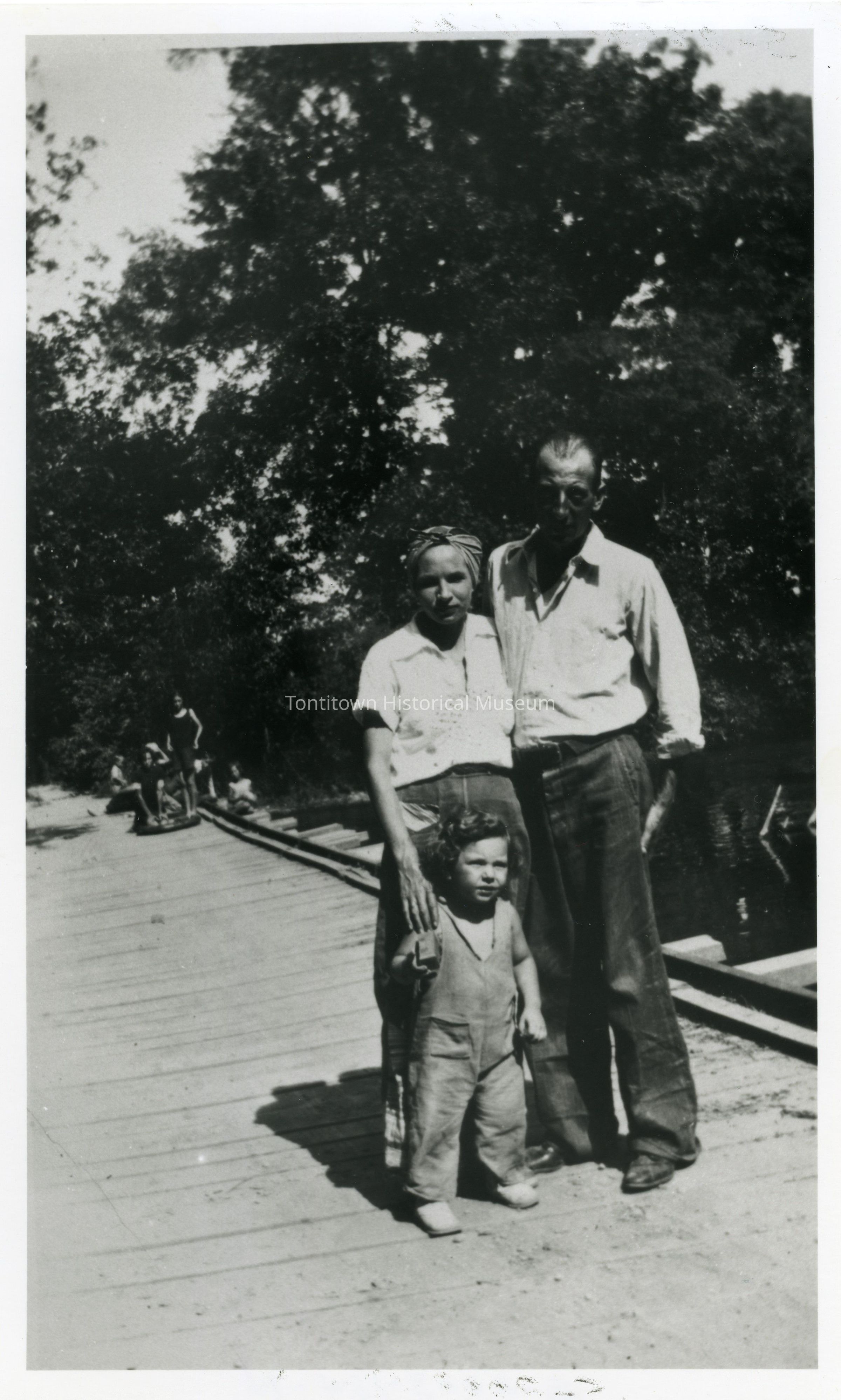 In this black and white photograph, three people—a man, a woman, and a child—stand closely together on a wooden pathway in what appears to be a rural setting with trees in the background, their faces relaxed and expressions serene. The man is wearing a light-colored shirt and dark pants, standing to the right with his arm gently resting on the woman’s shoulder, who is dressed in a short-sleeved blouse and a patterned headscarf, while the young child stands in front, wearing