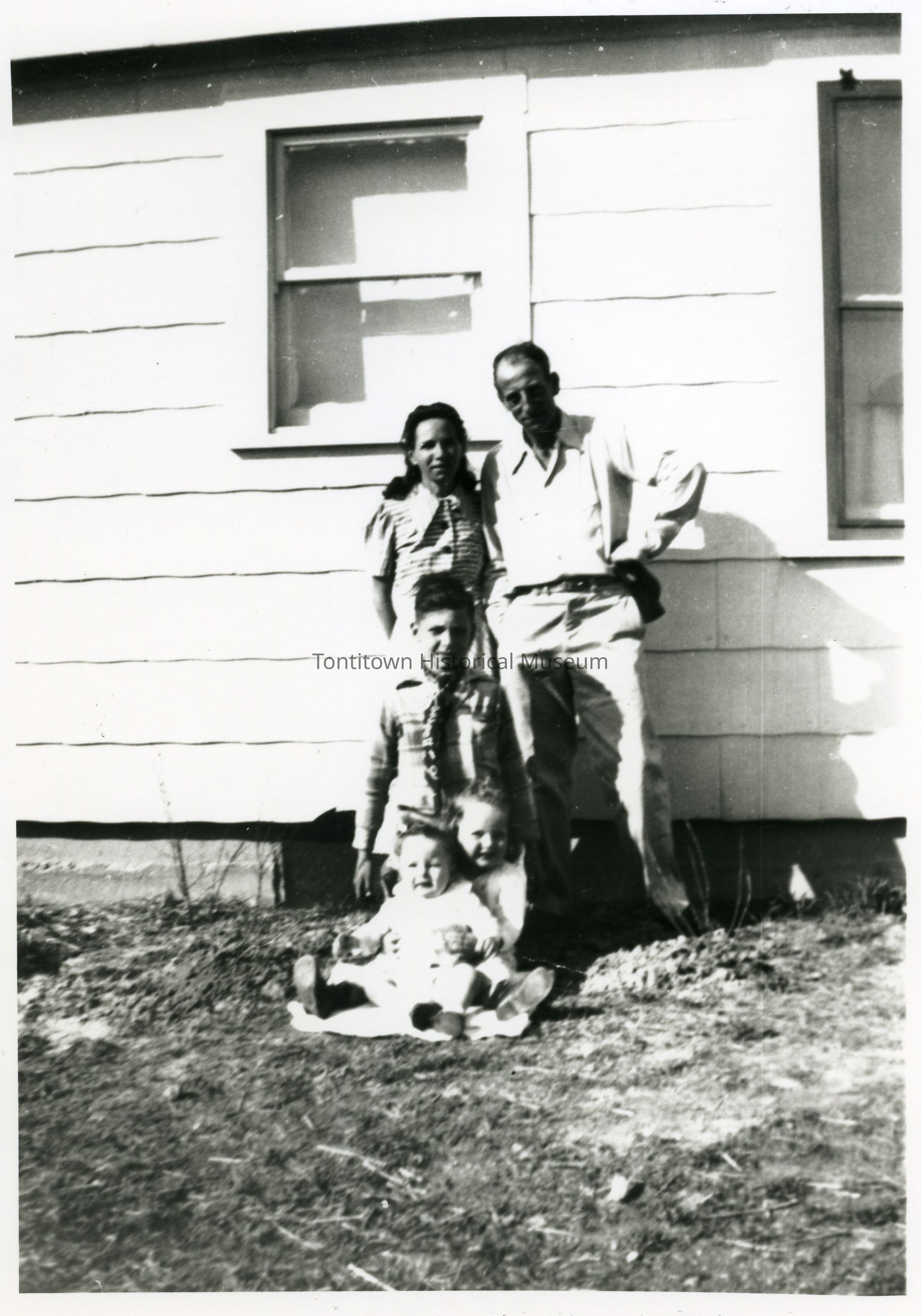 This black-and-white photograph captures a family of four posing in front of a modest clapboard house, with two adults standing side by side, seemingly the mother and father, displaying a relaxed posture and possibly wearing clothes typical of the early to mid-20th century; in front of them stands a young boy, likely a sibling, with a straight posture and a slight smile, while two younger children, possibly siblings as well, sit on the grass in front, displaying cheerful expressions and cozy body language;