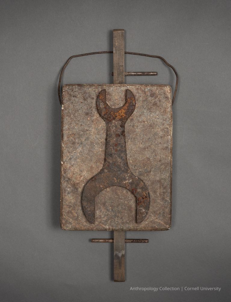 ICON OF STILT-WALKER | circa 1966-71 | Photo by Ann Cady, 2024 | ©2026 Norman Daly Art — This artifact is comprised of a rectangular, textured stone slab featuring a affixed shape resembling a modern-day wrench, which is mounted on a top-to-bottom wooden support; the stone’s surface appears weathered with patches of brown and gray, while the metal elements are rustic.
