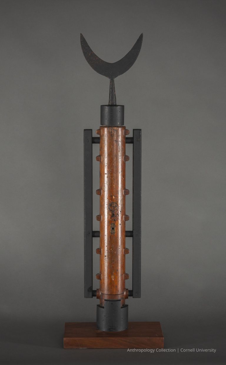 MACE | circa 1961-65 | Photo by Ann Cady, 2024 | ©2026 Norman Daly Art — Avertical metallic structure with a central cylindrical component held by two parallel handle-like black beams, topped by a crescent-shaped finial. It is displayed on a wooden base against a formal gray background.