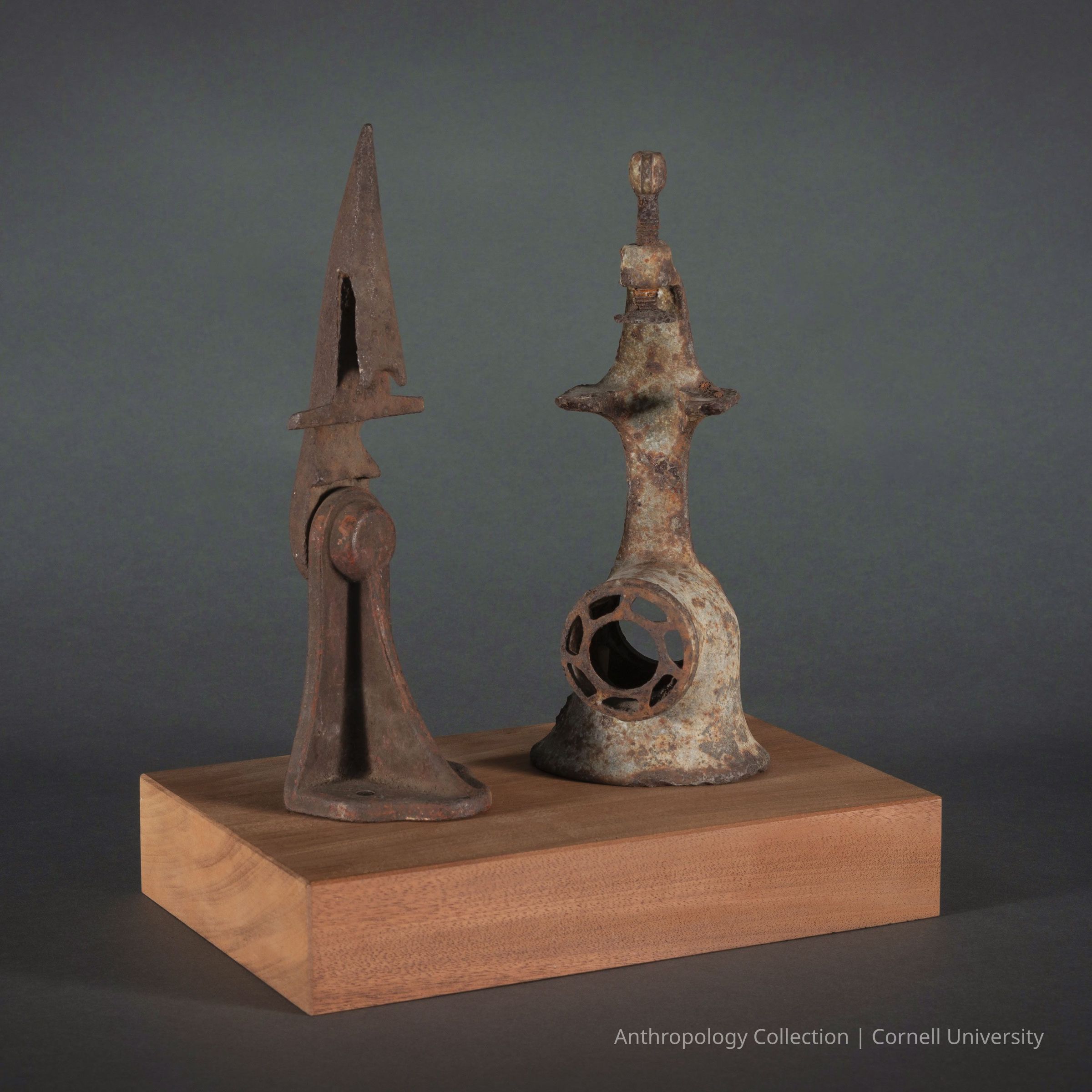 PAIR OF MINOR DEITIES | circa 1966-71 | Photo by Ann Cady, 2024 | ©2026 Norman Daly Art: Two figures made of rusted metal, each with unique shapes and textures, mounted on a smooth wooden base. The figure on the left is a tall, pointed structure with a triangular blade-like element, while the one on the right has a more cylindrical form with an open circle at the base and a small knob at the top. Their surfaces are rough and weathered, highlighting their age.