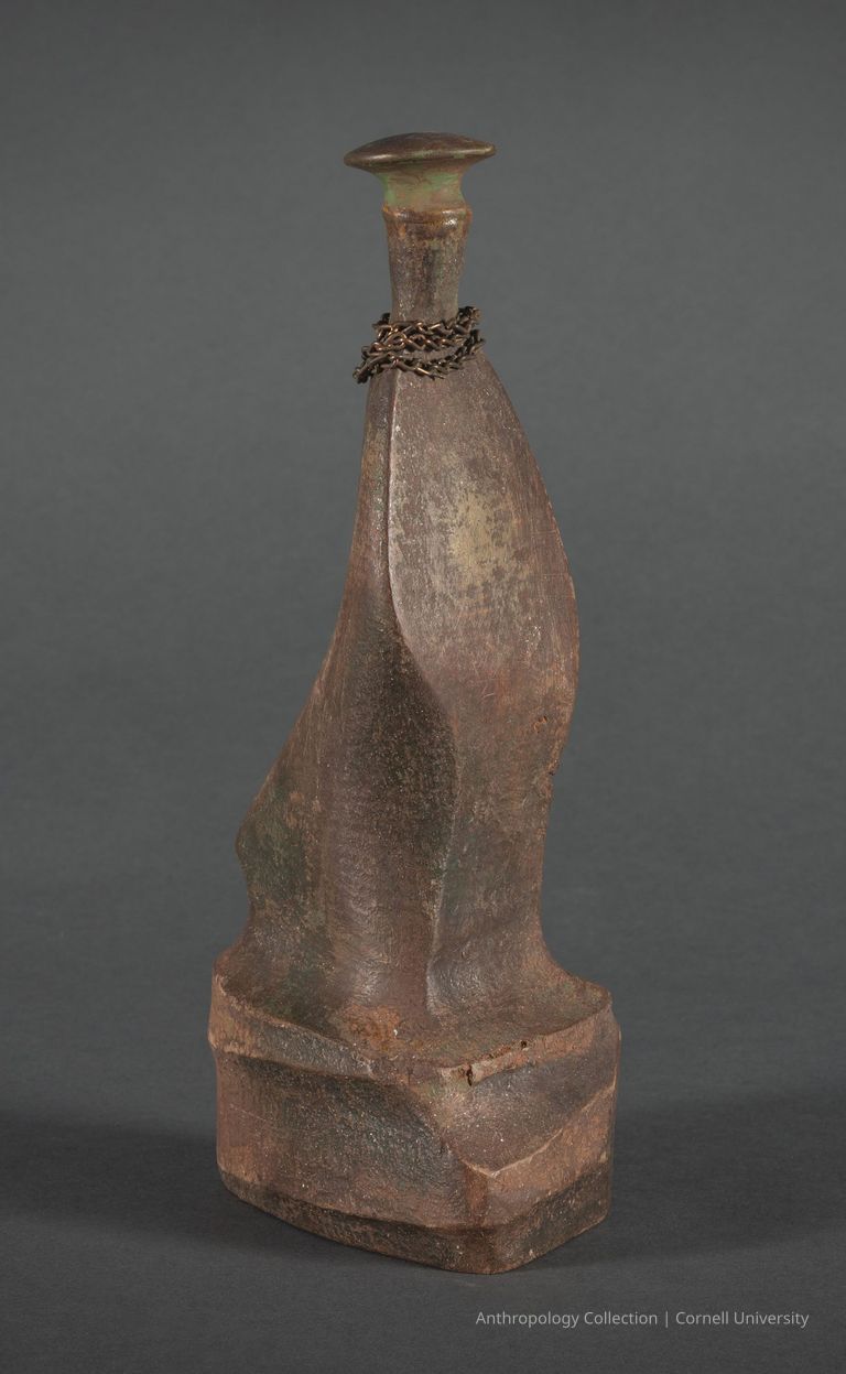 VOTIVE. PENITENT FIGURE | circa 1966-71 | Photo by Ann Cady, 2024 | ©2026 Norman Daly Art — An intricately designed, aged wooden artifact with a greenish and brown patina, featuring a uniquely curved and textured body, and topped with a wide, rounded 'stopper' reminiscent of a beret; a rusted chain is wrapped around the neck of the figure, enhancing its narrative mystery.