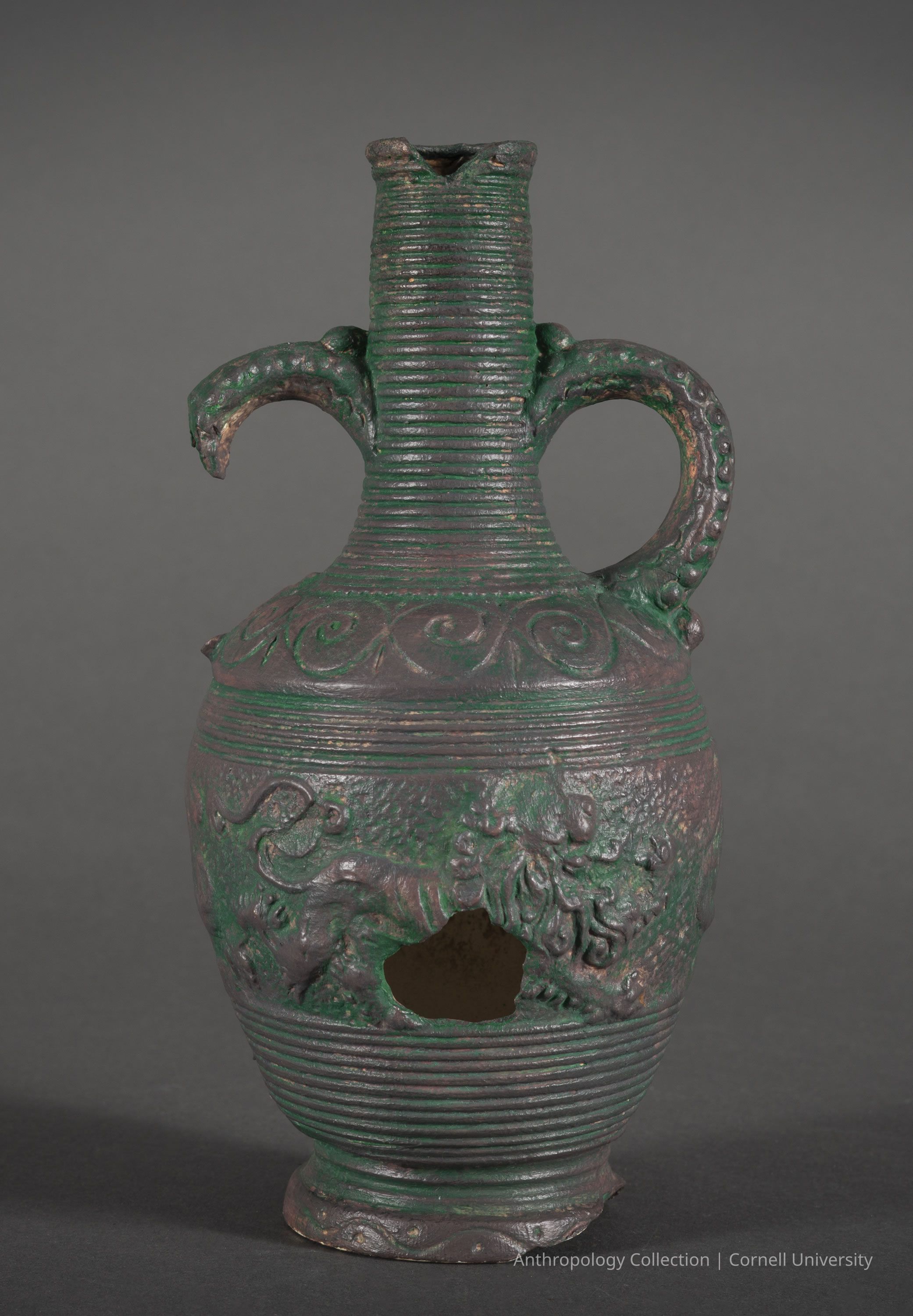 LOXOS | circa 1966-71 | Photo by Ann Cady, 2024 | ©2026 Norman Daly Art: A seemingly ancient, ornate bronze vessel with a green patina, featuring a narrow, ridged neck and two curved, intricately designed handles adorned with detailed embellishments. The body of the vessel showcases decorative swirling patterns near the top, and the lower part is adorned with elaborate relief carvings that appear to depict animals or creatures. Notably, there is a significant hole on the side of the vessel and chip in the lip which adds to its aged, historical appearance.