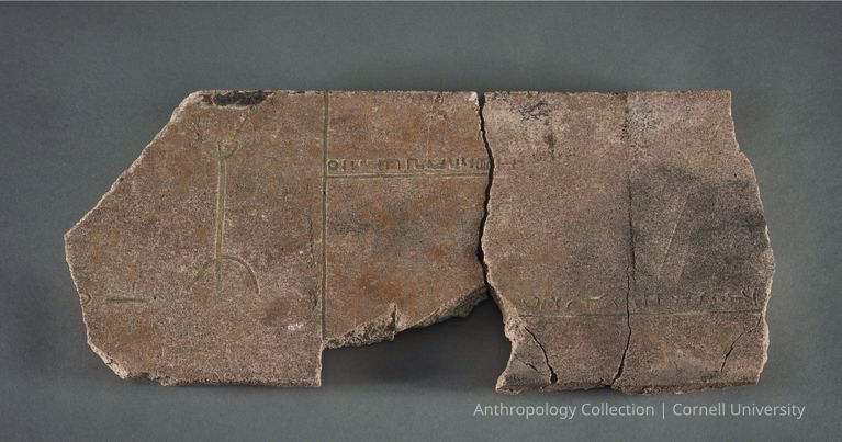 PETROGLYPH WITH STILT-WALKER | circa 1966-71 | Photo by Ann Cady, 2024 | ©2026 Norman Daly Art — A fragmented, weathered stone tablet featuring ancient inscriptions with faint and irregular markings, its surface cracked in places, on a flat dark surface, hinting at its historical significance and age. It  appears to depict a crude image of a stilt walker and Llhuroscian cuneiform.