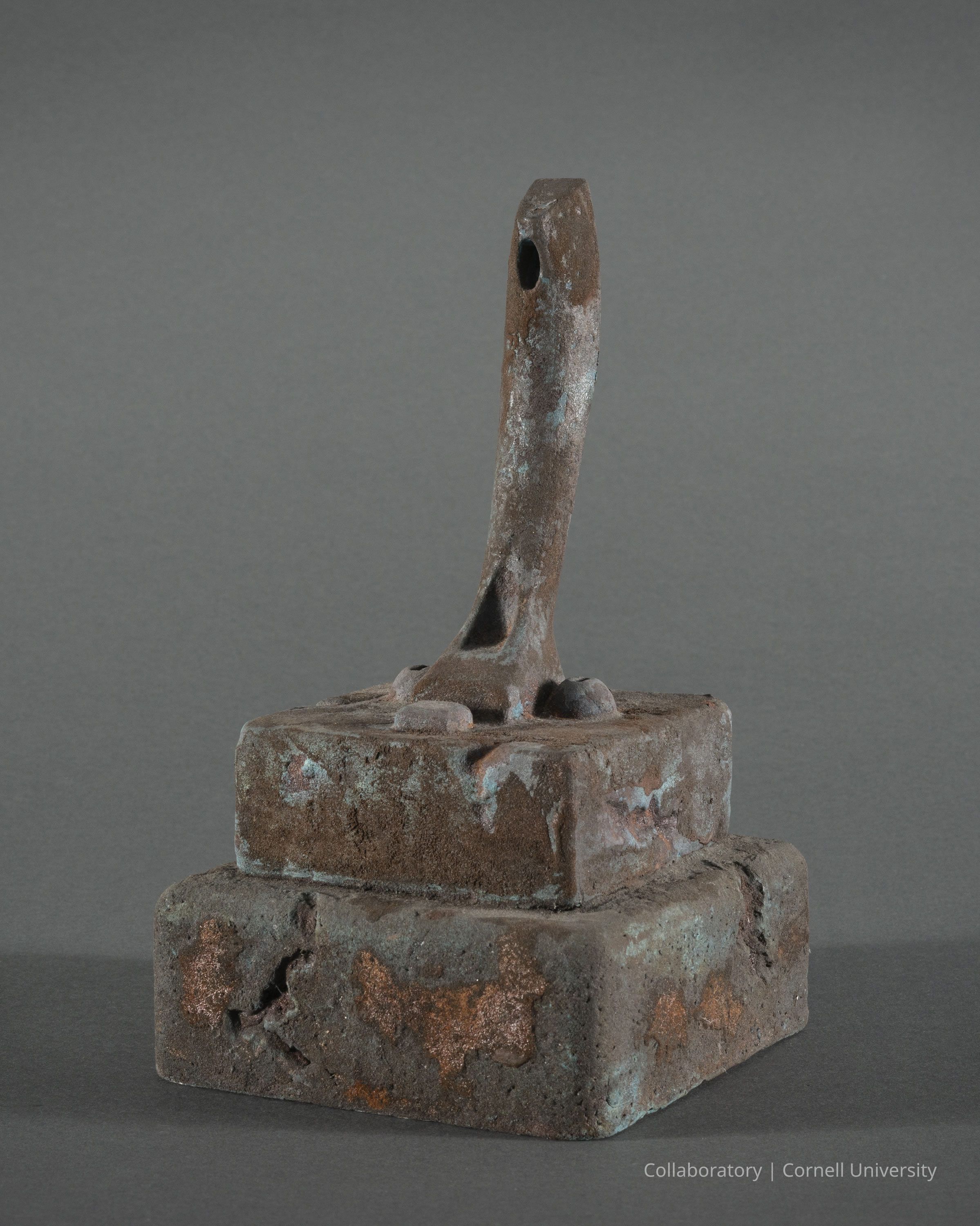 TEMPLE VOTIVE | circa 1974-1983 | Photo by Ann Cady, 2024 | ©2026 Norman Daly Art: A rustic, weathered, double-pedestal form with an erect phallic shape on top, exhibiting signs of rust and oxidation; the top layer is somewhat smaller compared to the base, which rests on a flat surface; together they create a geometric form with clearly visible, irregular patches of corrosion, providing a textured appearance against the plain, neutral backdrop. The phallic shape suggests its use as a votive in a temple, perhaps the Temple of Phallus at Trolydos.