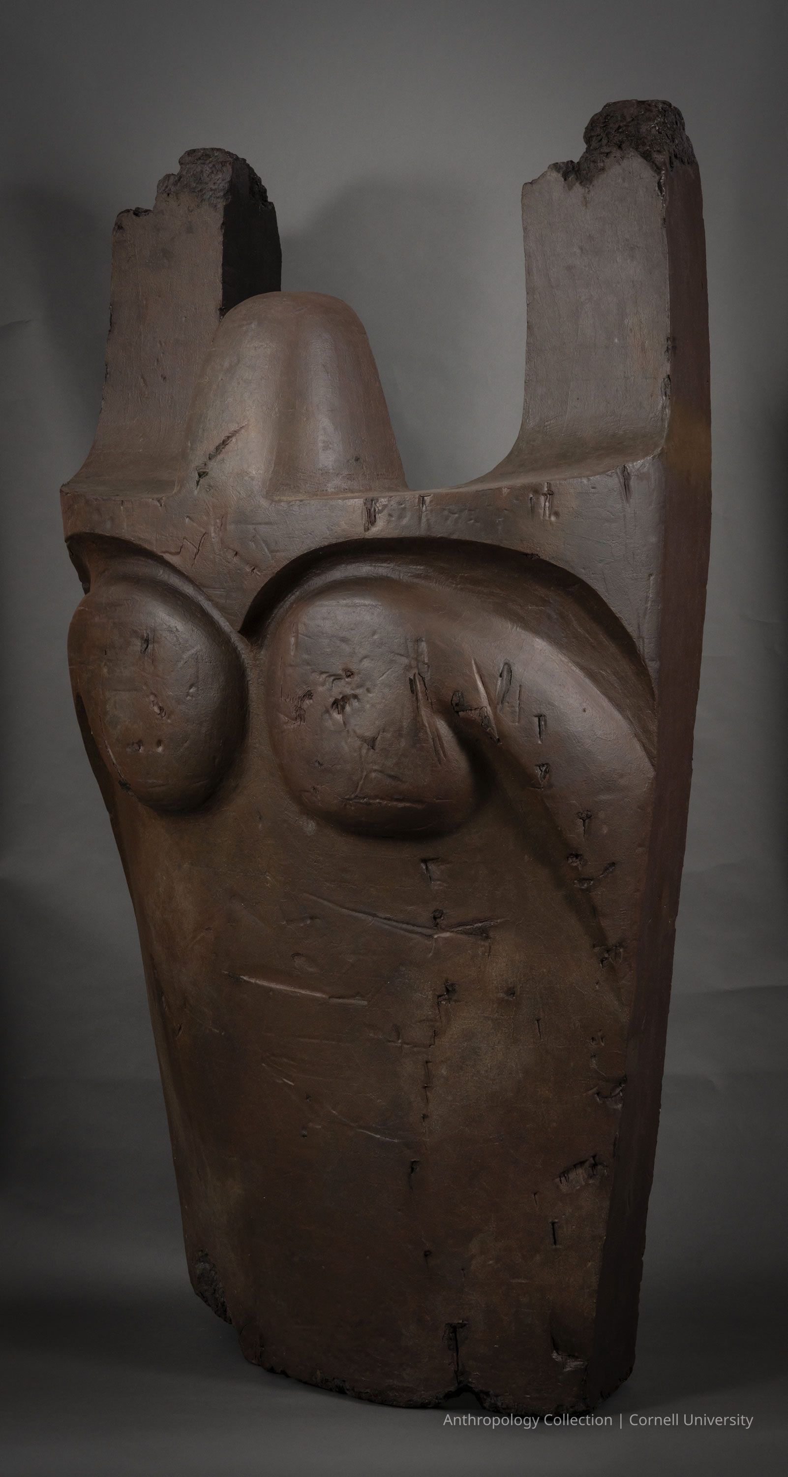 FRAGMENT OF THE EFFIGY OF HOLMEEK | circa 1966-7 1 Photo by Ann Cady, 2024 | ©2026 Norman Daly Art: One of the largest most impressive discoveries during the archaeological dig in Llhuros, this photo showcases a large, vertical, fragment of a figure made of bronze, thought to be the Llhuroscian god, Holmeek. The effigy appears ancient and weathered, with rough textures, cracks, and scratches. Its tall, narrow form culminates in a jagged, uneven top, indicating fracture or erosion.