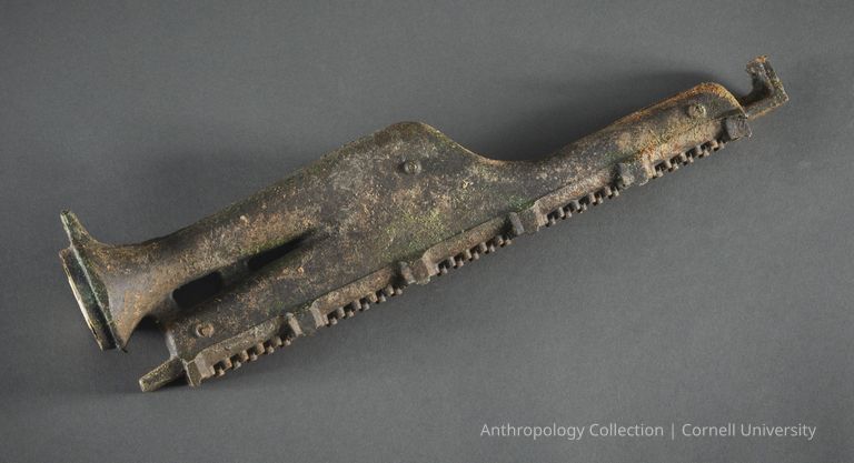 NASAL FLUTE | circa1966-71 | Photo by Ann Cady, 2024 | ©2026 Norman Daly Art — The photo features a weathered metal object with a distinct elongated shape, sturdy structure, and prominent, textured surface, showcasing signs of age and oxidation  the object includes intricate details such as toothed edges and a flared, trumpet-like end, indicating it may have served a musical function in its time; positioned against a formal, dark gray background.