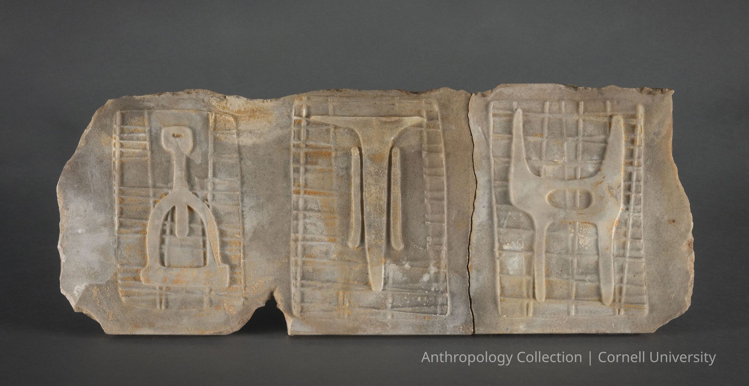 VOTIVE OF STILT-WALKERS | circa 1966-71 | Photo by Ann Cady, 2024 | ©2026 Norman Daly Art: A marble fragment, with each of three distinct carvings presenting symbolic figures over grid-like patterns. The reliefs appear weathered and fragmented, suggesting significant archaeological interest, possibly originating from skilled stone-carvers of Llhuros. The carvings include identifiable icons that could represent cultural or religious symbols, emphasizing their likely role in rituals or as markers of social or spiritual practices such as the rite of stilt-walking.