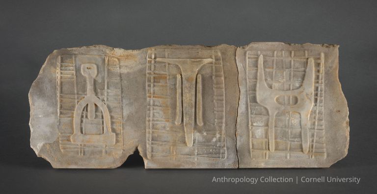 VOTIVE OF STILT-WALKERS | circa 1966-71 | Photo by Ann Cady, 2024 | ©2026 Norman Daly Art — A marble fragment, with each of three distinct carvings presenting symbolic figures over grid-like patterns. The reliefs appear weathered and fragmented, suggesting significant archaeological interest, possibly originating from skilled stone-carvers of Llhuros. The carvings include identifiable icons that could represent cultural or religious symbols, emphasizing their likely role in rituals or as markers of social or spiritual practices such as the rite of stilt-walking.