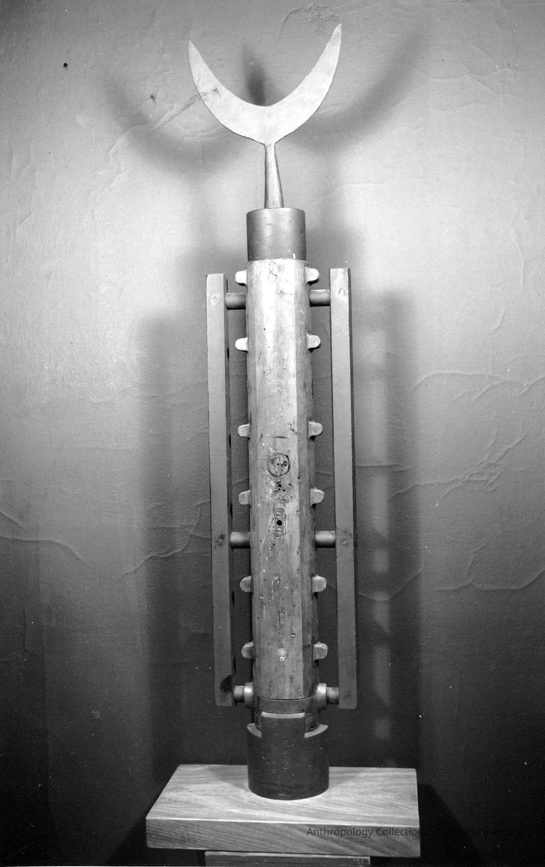 CONSTRUCTION | a.k.a COL.56 | circa 1961-65 | Photo by Marion Wesp | ©2026 Norman Daly Art — The black and white photo of previous object shows a wooden totem-like assemblage on pedestal with a metal crescent-shaped finial on top. Displayed with its shadow against a crudely plastered wall.