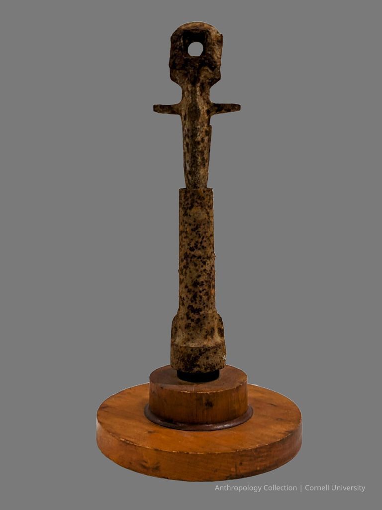 VOTIVE FIGURE | circa 1966-1971 | Photo by Linda Fisher, 2024 | ©2026 Norman Daly Art — A rustic iron artifact mounted on a circular wooden base, prominently displaying an elongated figure with arms extending outward and a circular head, overall suggesting a religious artifact possibly used in ritualistic practices or as a totemic object. The artifact's mottled texture and color indicate aging and exposure to the elements.