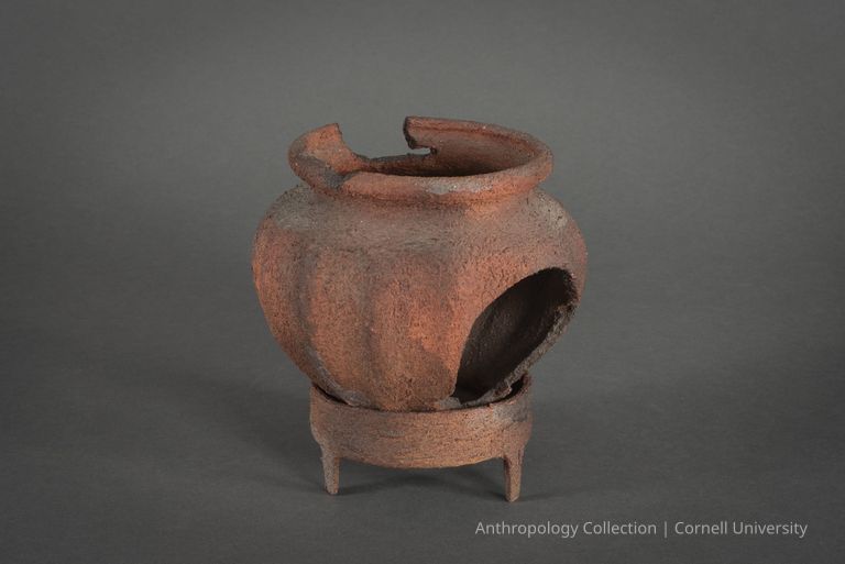 WATER CLOCK | 1966-71 | Photo by Ann Cady, 2024 | ©2026 Norman Daly Art — An ancient, weathered pottery urn with a rustic, orange-brown surface texture, resting on a three-legged stand. The vessel has significant signs of wear and aging, including a large, jagged opening on one side and a partially broken rim, revealing a dark interior. The artifact's textured and corroded surface, emphasizes its archaeological significance and the passage of time it has endured.
