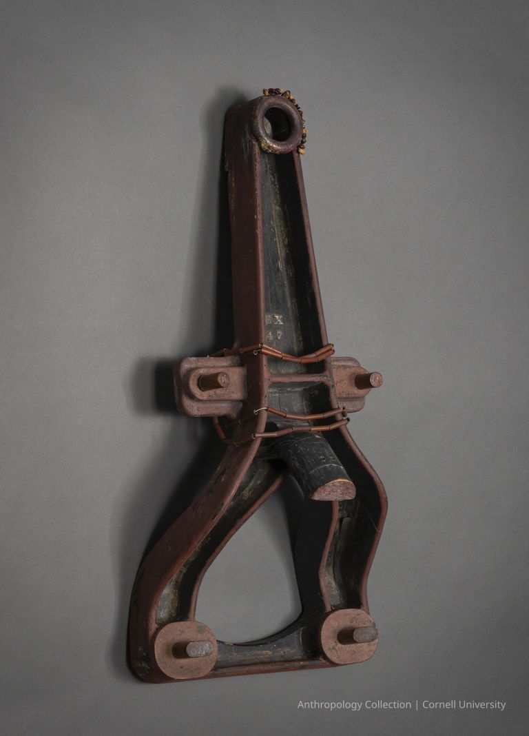 FUNEREAL HERO | circa 1966-71 | Photo by Ann Cady, 2024 | ©2026 Cornell University — A wooden object with a symmetrical design, featuring a central vertical element that tapers towards the top, where a circular opening is present, while the lower portion has two curved legs, suggesting the appearance of a triangular frame or support structure; the object has a rustic, aged appearance with visible wear, displaying different shades of brown and black, decorated with beads; hinting at its possible functional purpose in a historical or cultural context.