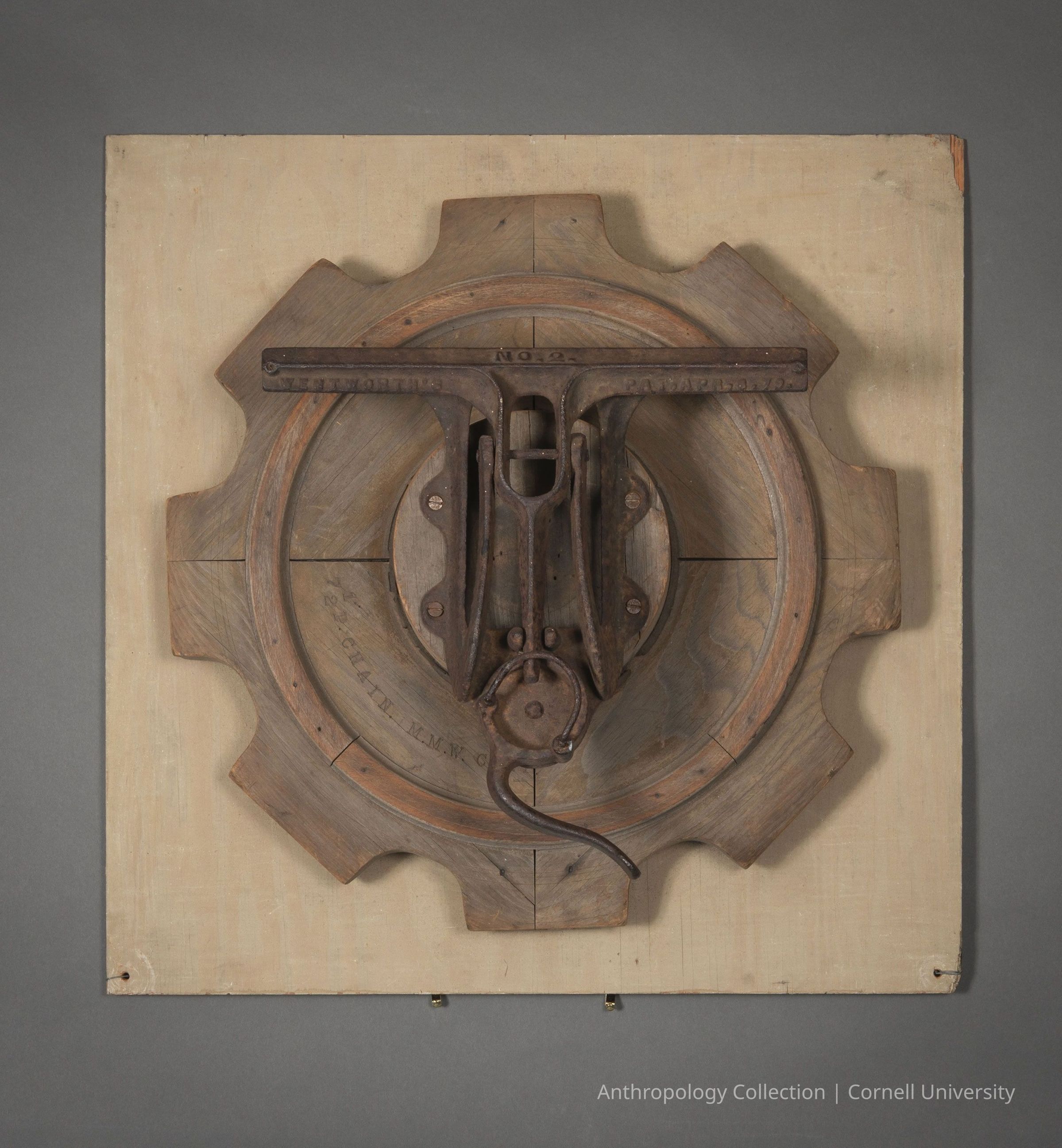 ASTROLABE | circa 1966-71 | Photo by Ann Cady, 2024 | ©2026 Norman Daly Art: The photo displays a wooden assemblage that appears to be a piece of industrial machinery, possibly a gear or cog-like design, mounted on a rectangular, flat wooden panel. The central part is constructed using darker wood, showcasing intricate craftsmanship with a circular pattern in the middle and a prominent overhanging metal piece, suggesting a mechanical component.