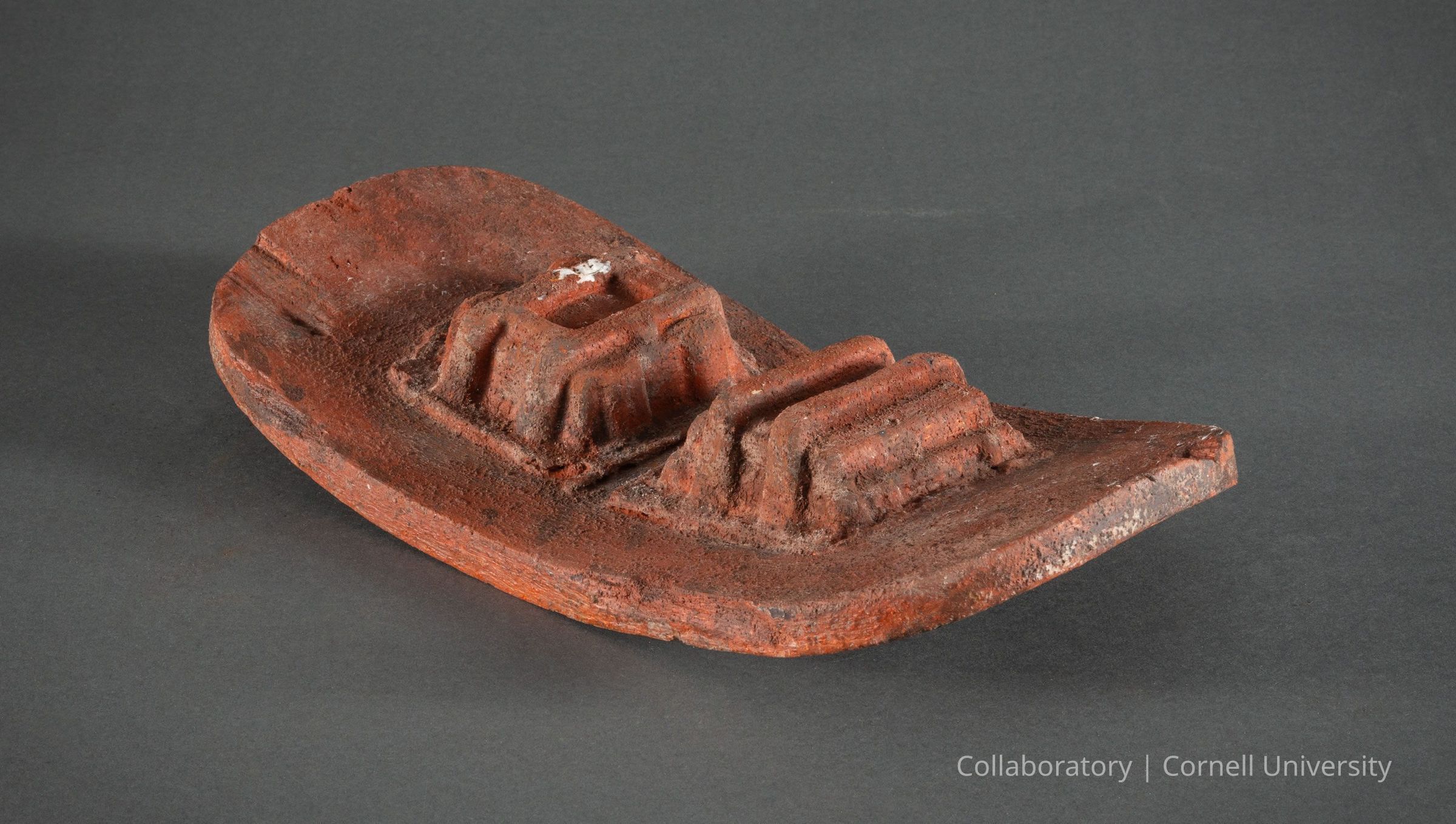 BIRD ROOST| 1966-71 | Photo by Ann Cady, 2024 | ©2026 Norman Daly Art: The photo is of a curved terracotta-colored artifact placed against a neutral gray background. It is thought to be a bird roost worn on the head of a stilt-walker during a pilgrimage. The roost has a rough, textured surface with two rectangular protrusions on top, suggesting perches where a bird was able to keep a foothold. The piece appears aged, with signs of wear and slight imperfections consistent with an ancient or handcrafted object.